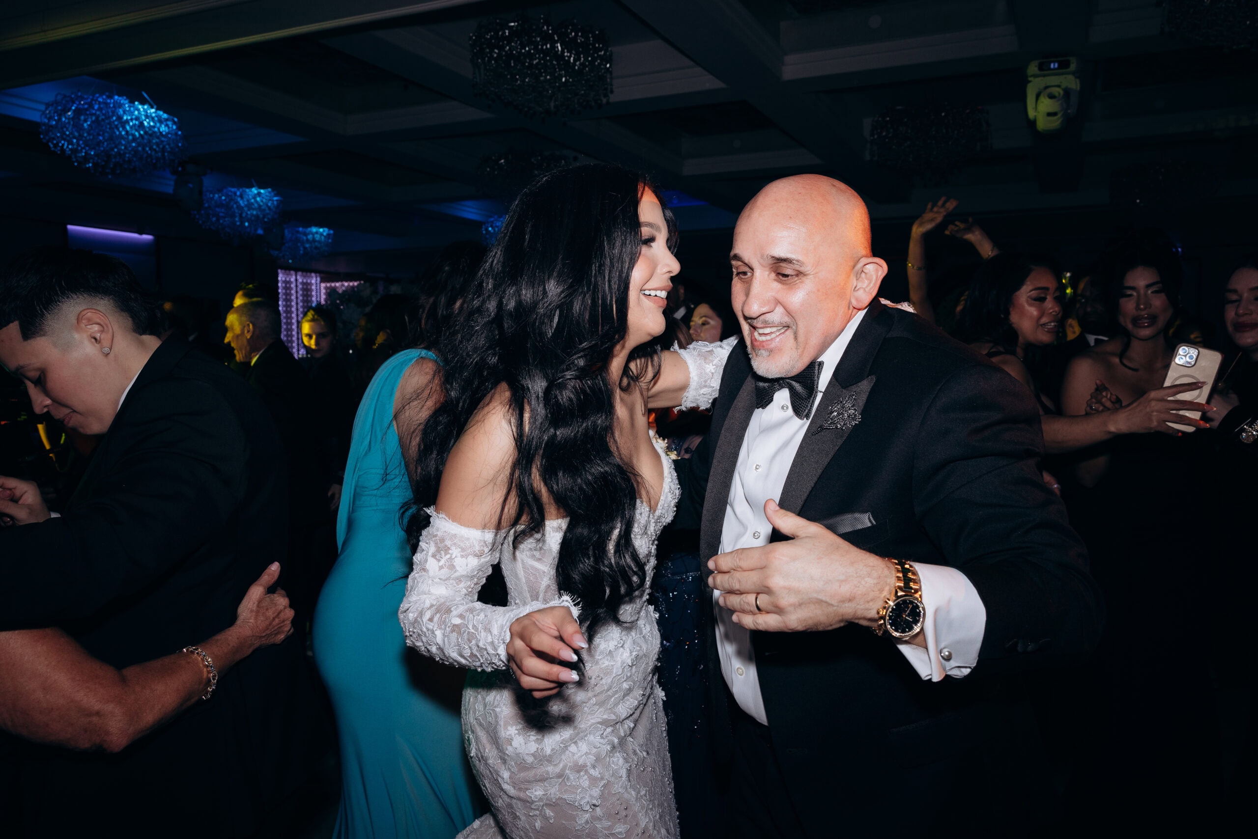 Bride in detailed lace wedding dress dancing closely with an elder man in black tuxedo and bow tie at a dimly lit wedding reception.