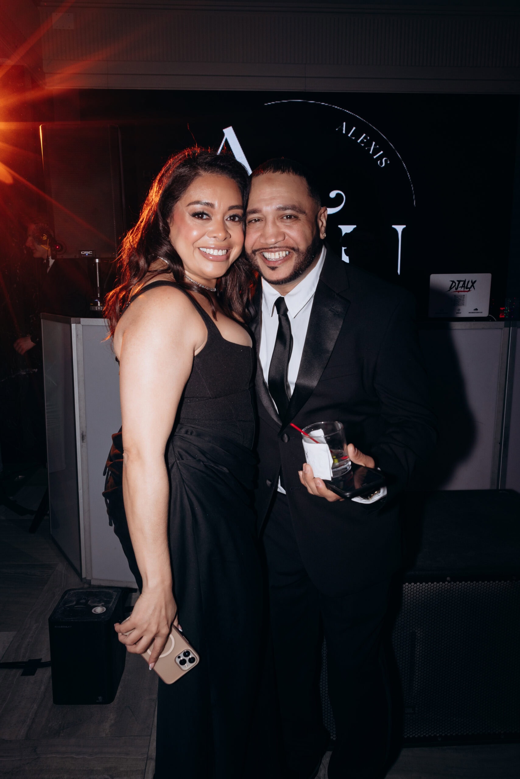 Man and woman in black formal attire smiling closely together inside an event space with warm lighting and a DJ booth in the background.