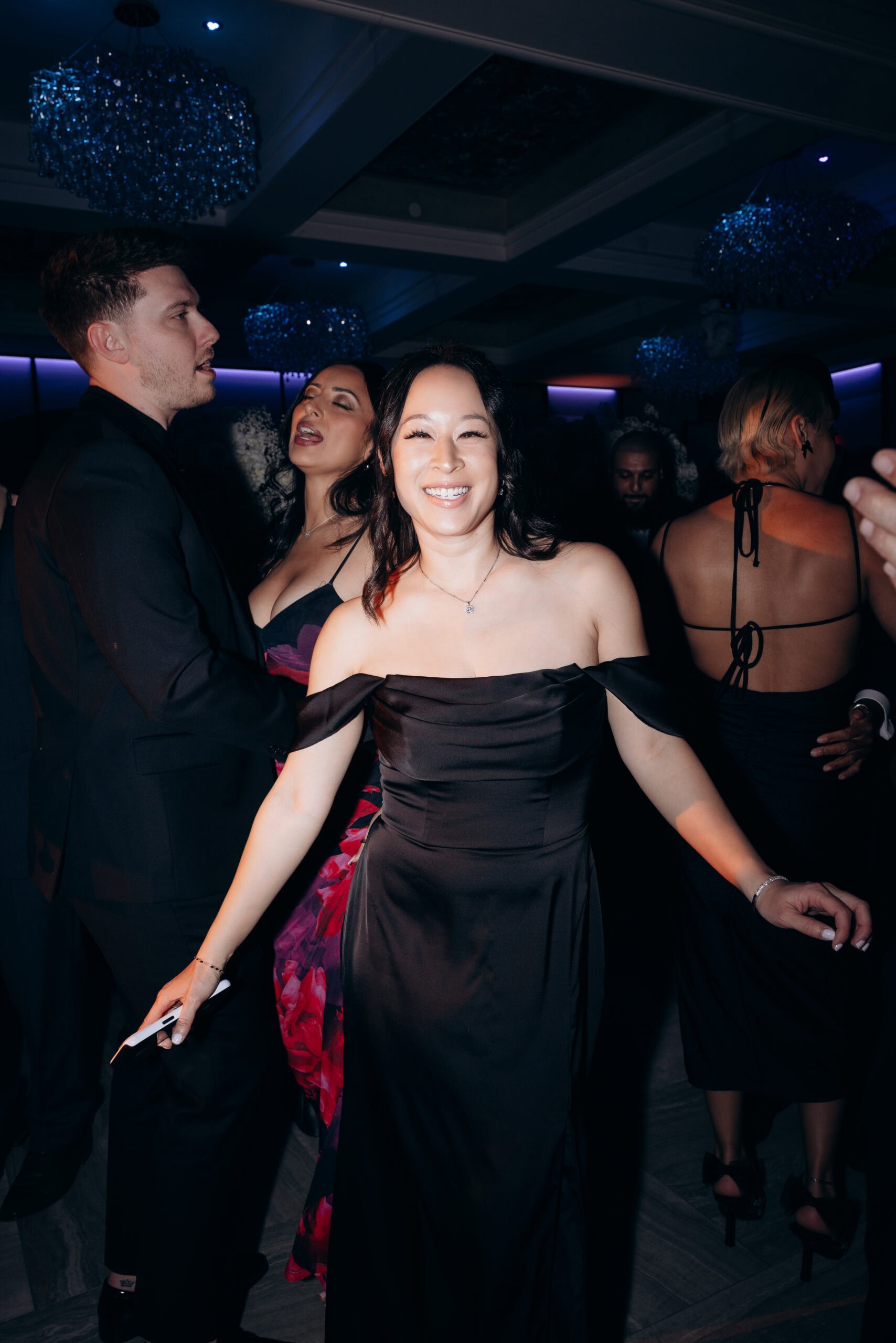 Woman in off-shoulder black dress smiling at the camera, surrounded by guests in formal black attire dancing under crystal chandeliers in a dimly lit venue.