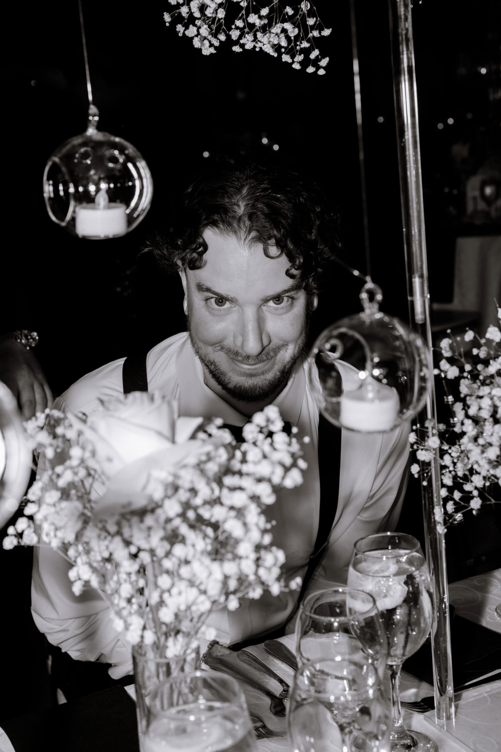 Close-up of a man with curly hair and a beard smiling subtly behind white roses and baby's breath flowers, surrounded by hanging glass candle holders at a wedding reception.