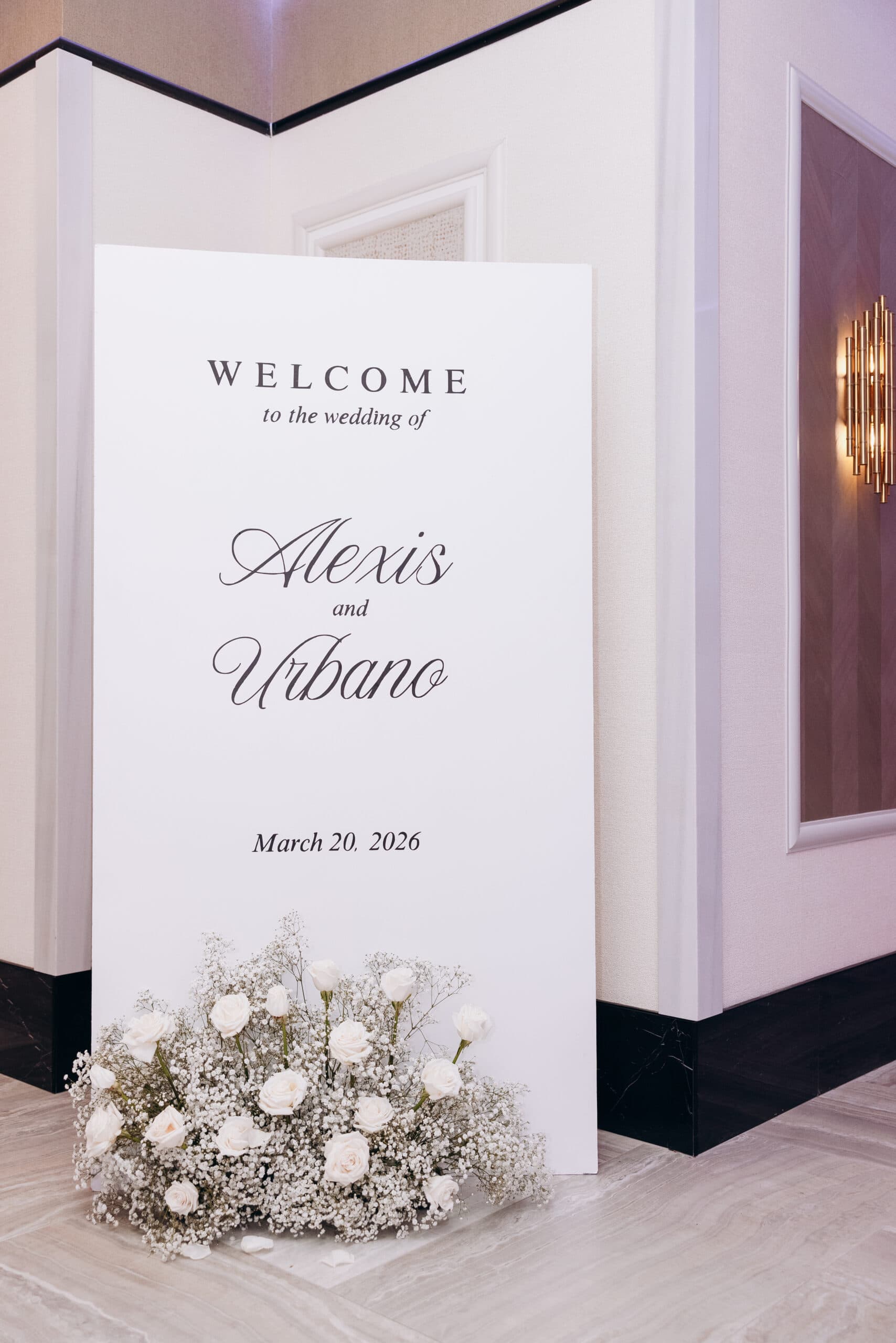 White wedding welcome sign with elegant black script reading Alexis and Urbano, March 20, 2026, adorned with white roses and baby's breath flowers at The Grand venue.