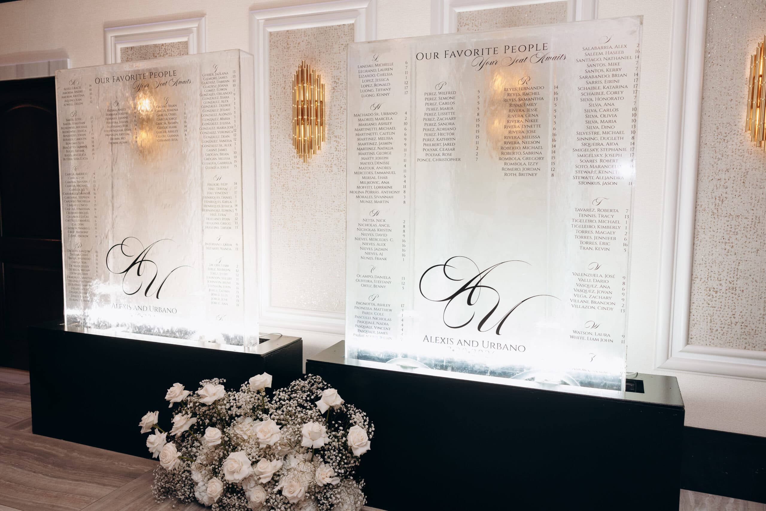 Two illuminated acrylic wedding seating charts with elegant calligraphy listing guest names, placed on black stands with white roses and baby's breath flowers in front.