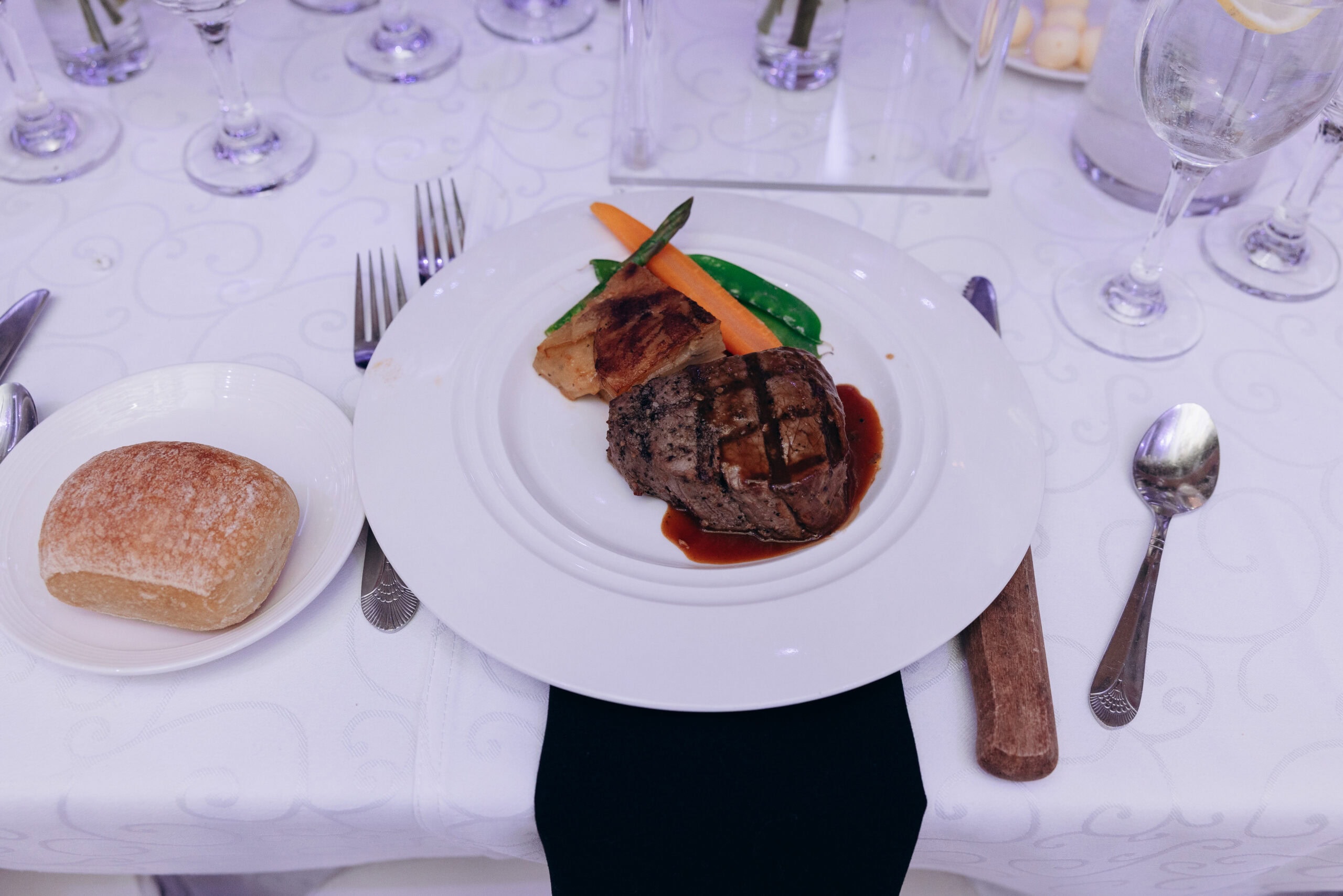 Plated grilled filet mignon with baby carrot, snap peas, and potato gratin on white plate, rustic bread roll on side plate, set on white embroidered tablecloth with glassware and cutlery.