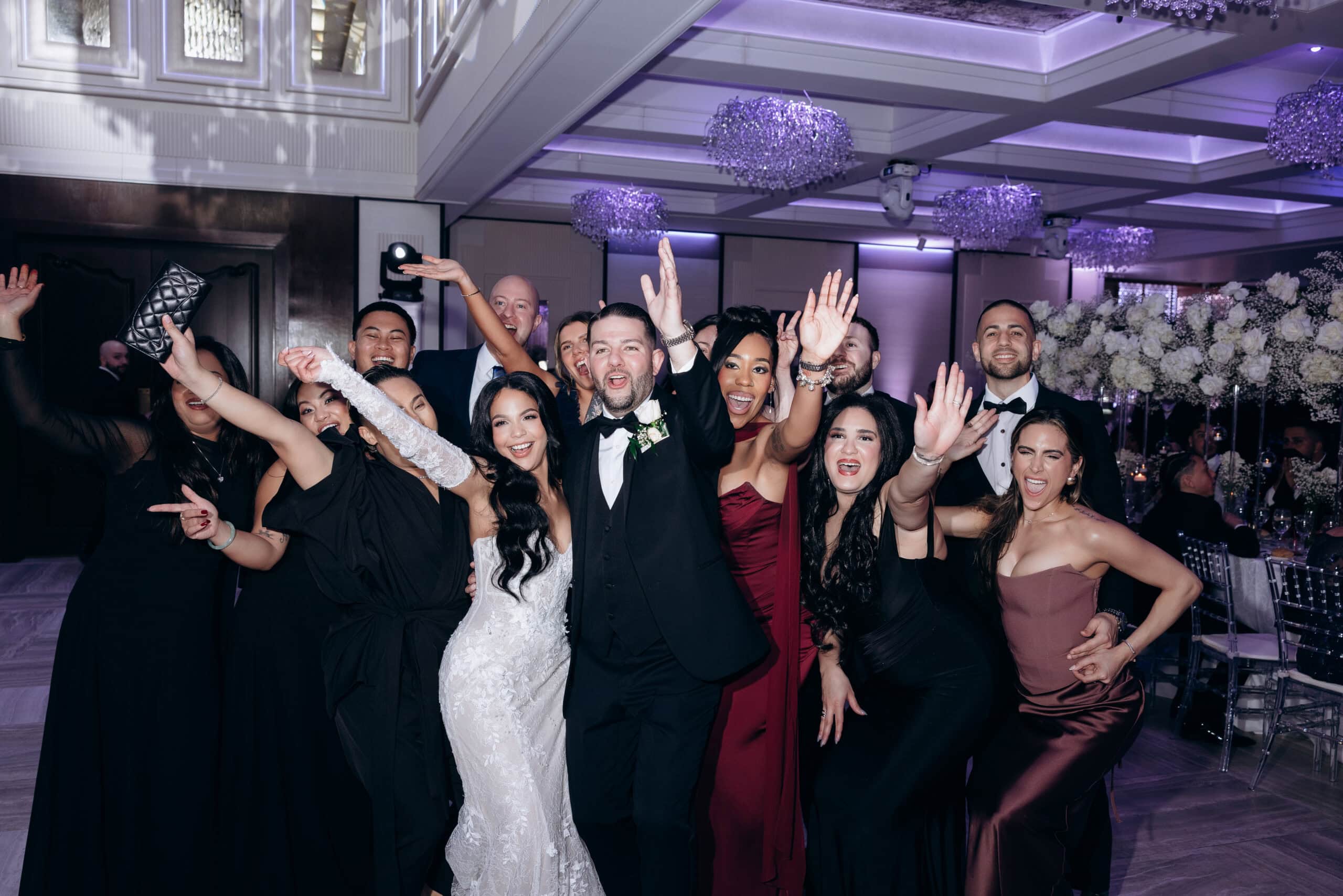 Wedding guests in formal attire cheer and wave in a lavishly decorated ballroom with purple lighting and floral arrangements at The Grand.