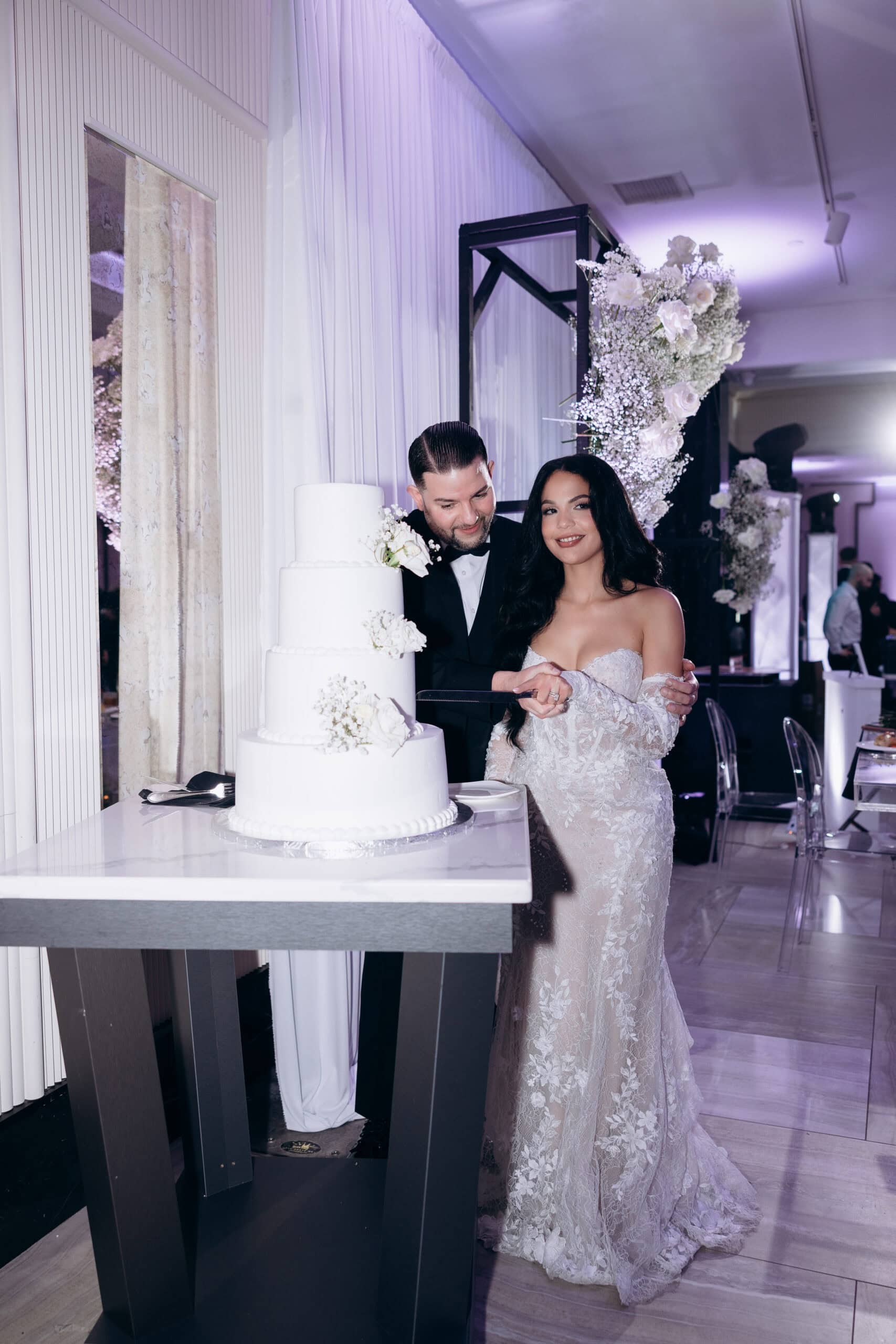 Bride and groom cutting a four-tier white wedding cake decorated with white flowers, standing in a softly lit venue with purple uplighting and floral decor.