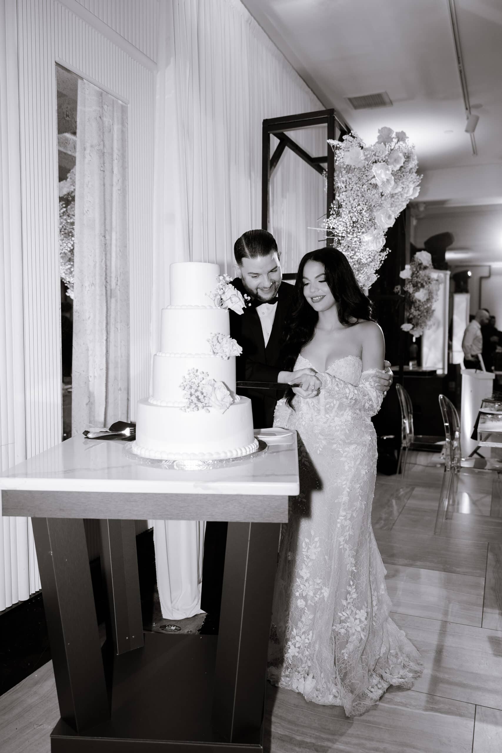 Bride and groom cutting a four-tier white wedding cake decorated with white roses and baby's breath at an indoor wedding reception with lavender lighting and floral decor.
