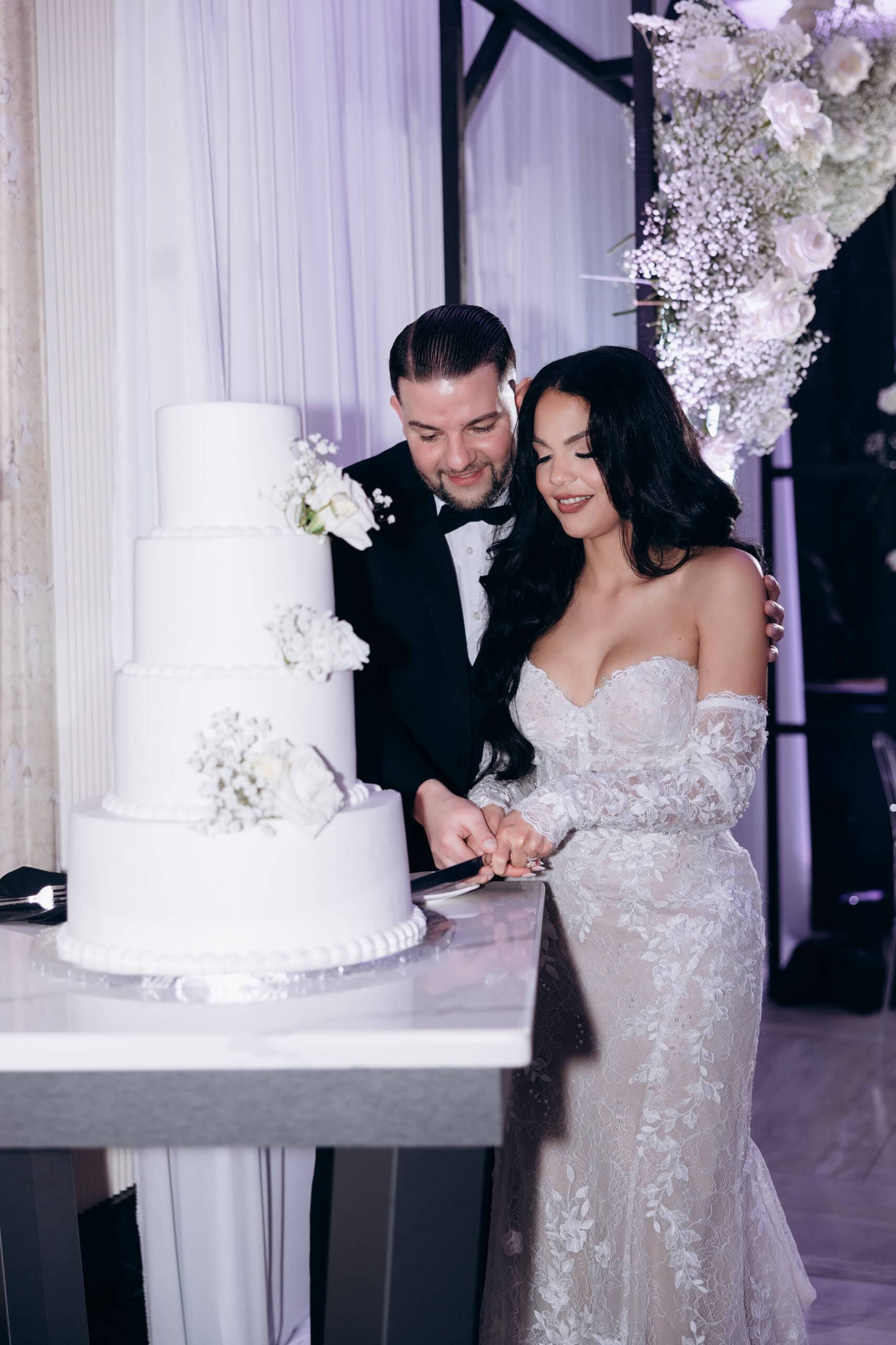 Bride and groom cutting a four-tier white wedding cake decorated with white roses and baby's breath, surrounded by elegant floral arrangements and soft curtains at The Grand.
