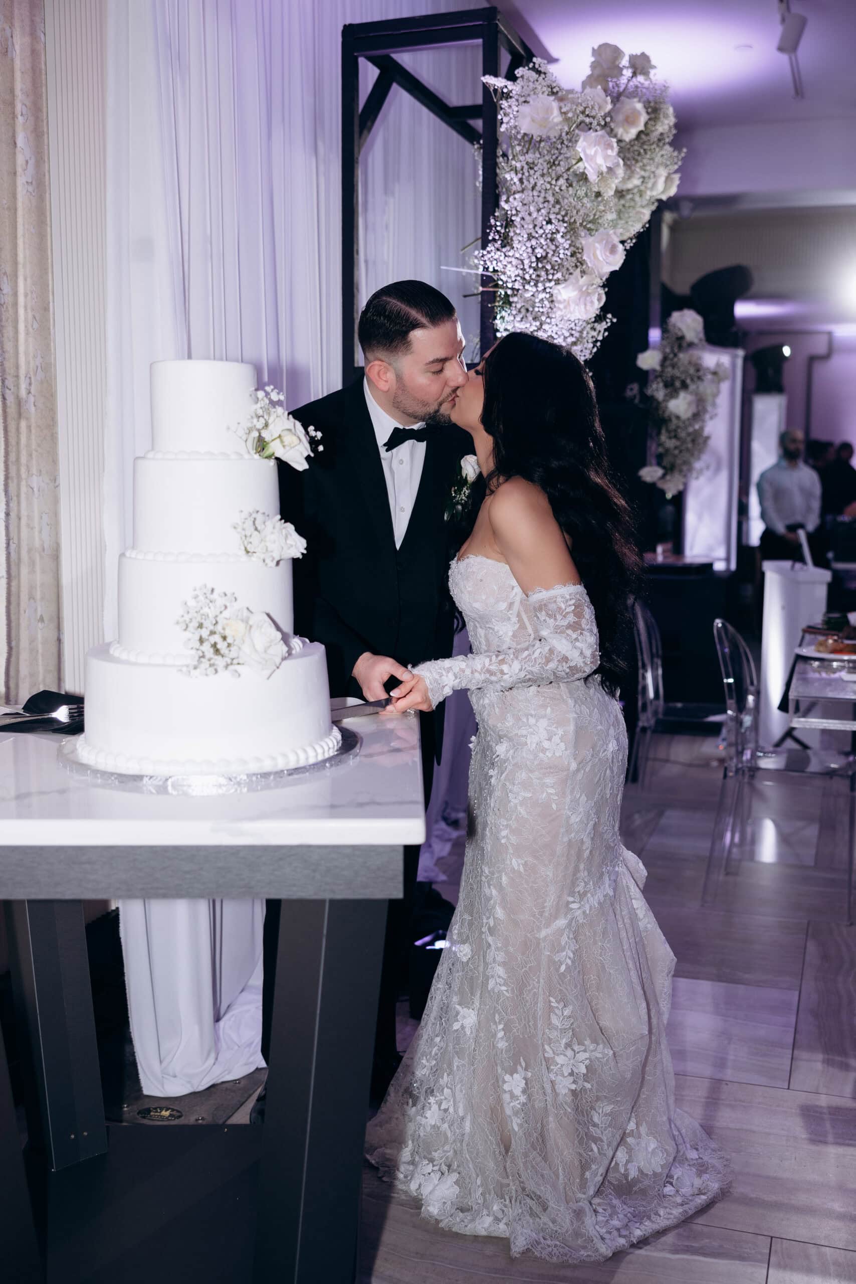 Bride and groom kissing while cutting a four-tier white wedding cake decorated with white roses and baby's breath, with floral arrangements and purple lighting in the background at The Grand.