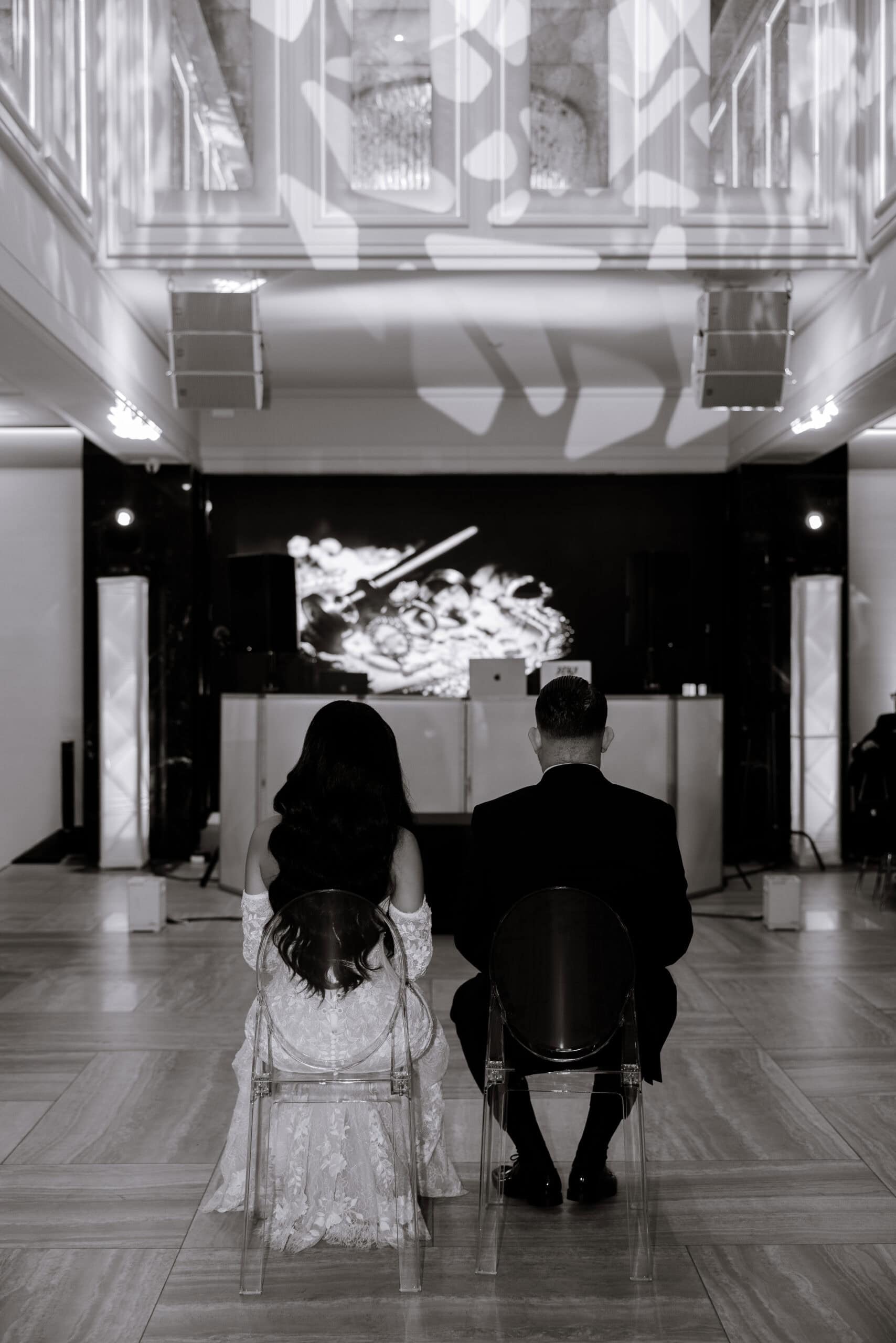 Rear view of a couple seated on clear chairs in a modern venue with abstract light projections on the ceiling and a softly lit stage in front of them.