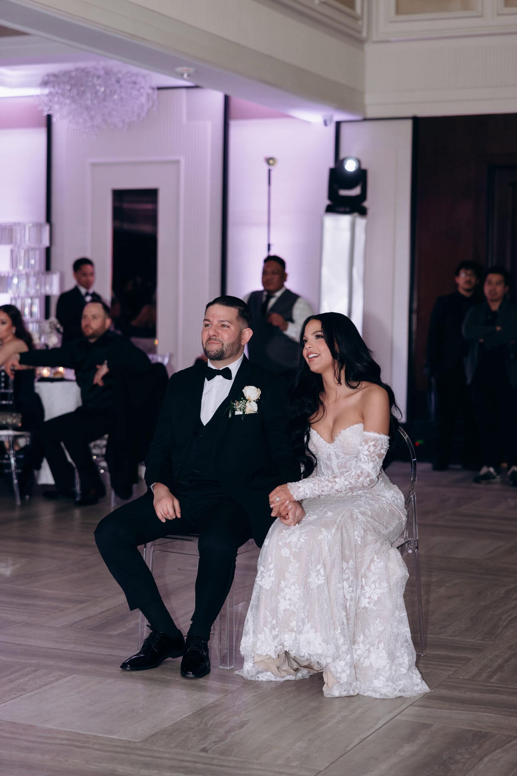 Bride in lace off-shoulder gown and groom in black tuxedo seated holding hands during wedding reception with purple uplighting at The Grand venue.