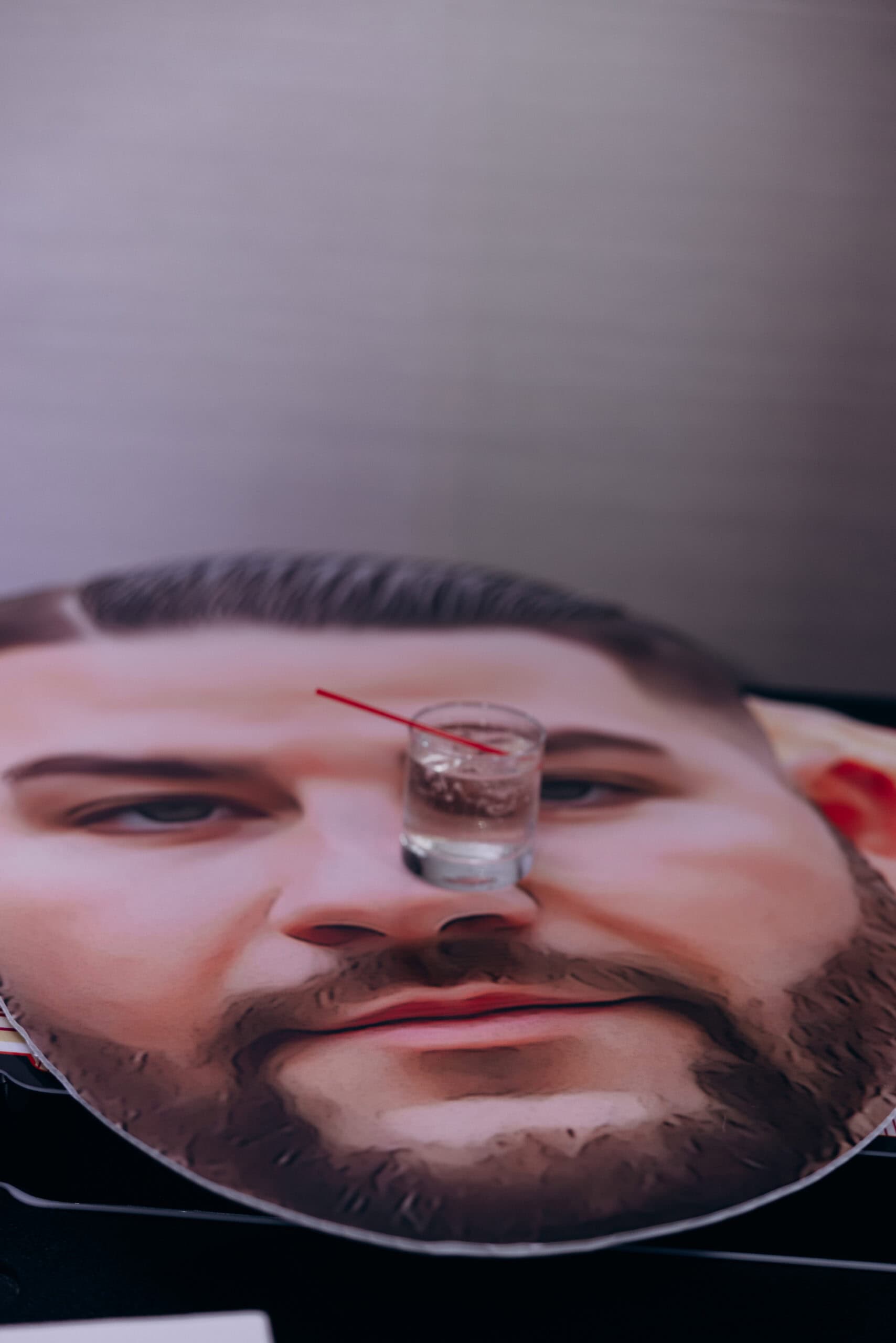 Close-up of a table with a large printed portrait of a man's face, with a clear glass of water and red straw placed on the nose area.