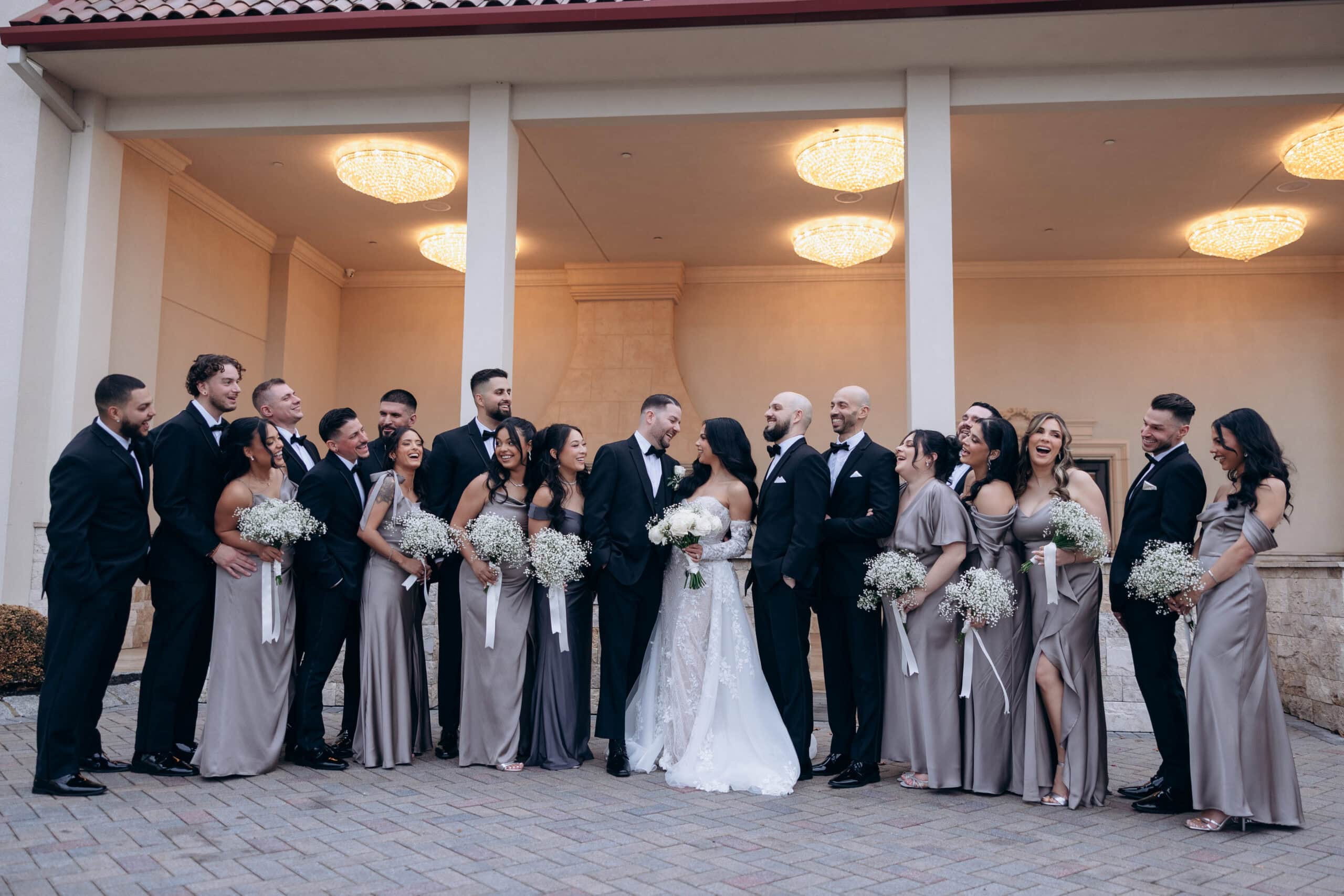 Wedding party in coordinated taupe dresses and black tuxedos standing under illuminated chandeliers at The Grand venue.