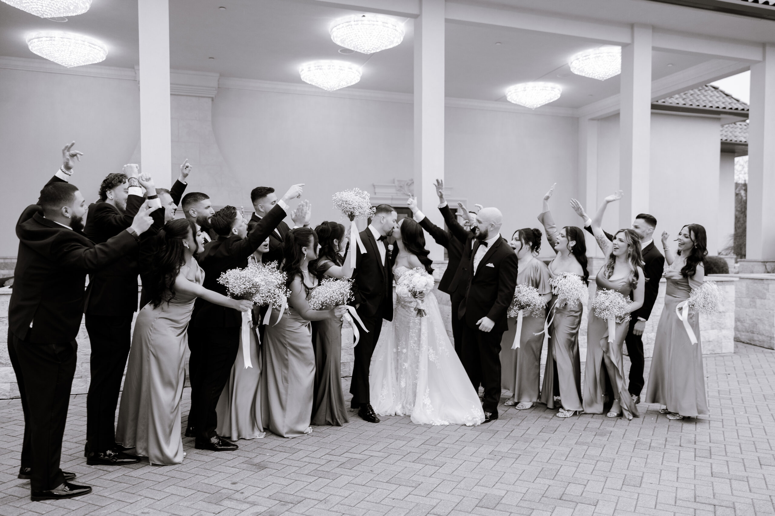 Black and white photo of a wedding party at The Grand, with bridesmaids in satin dresses holding bouquets and groomsmen in tuxedos surrounding the bride and groom sharing a kiss.