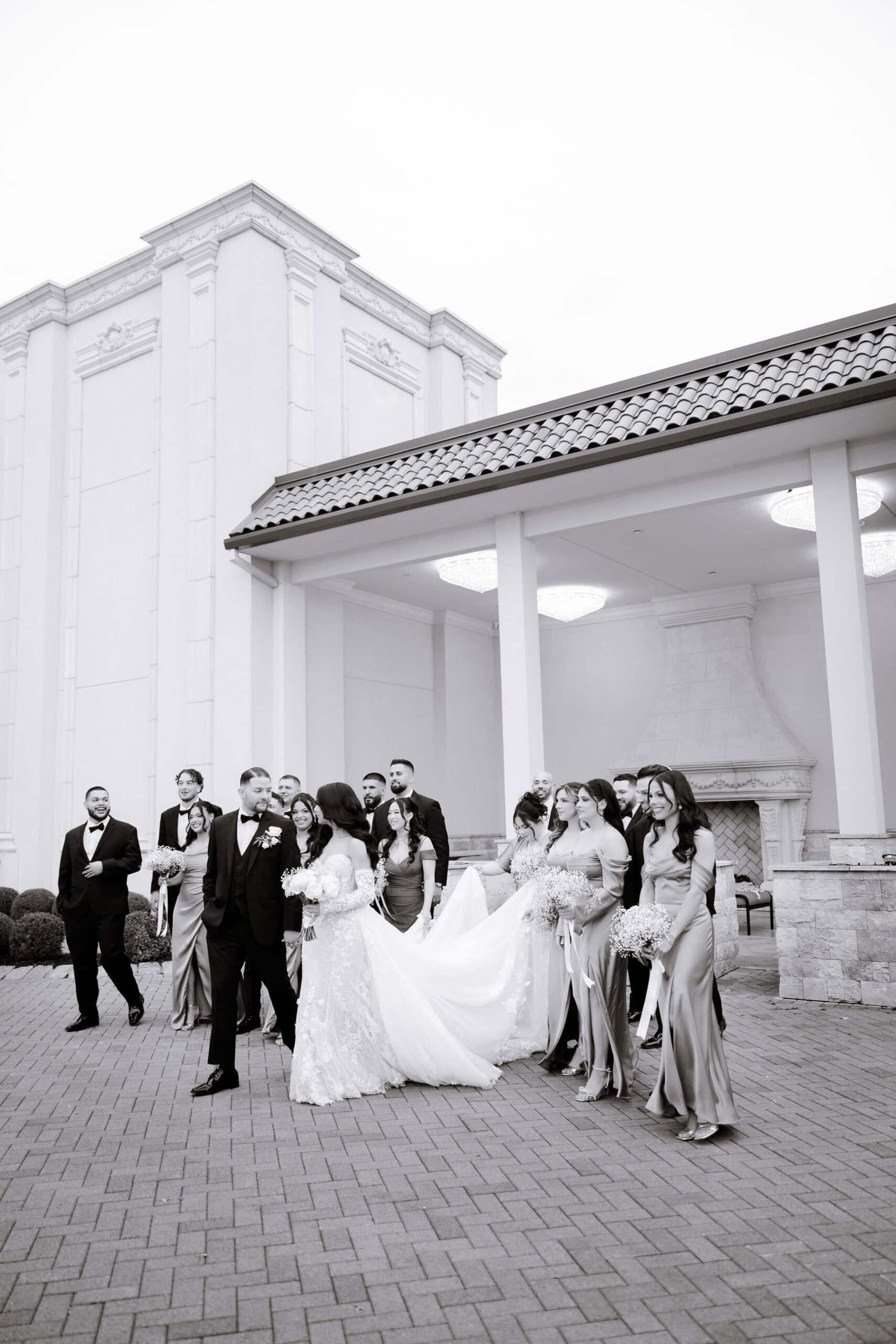 Black and white photo of a wedding party dressed in formal attire standing outside a grand building with classical architecture, featuring the bride in a lace gown and bridesmaids in satin dresses holding bouquets.
