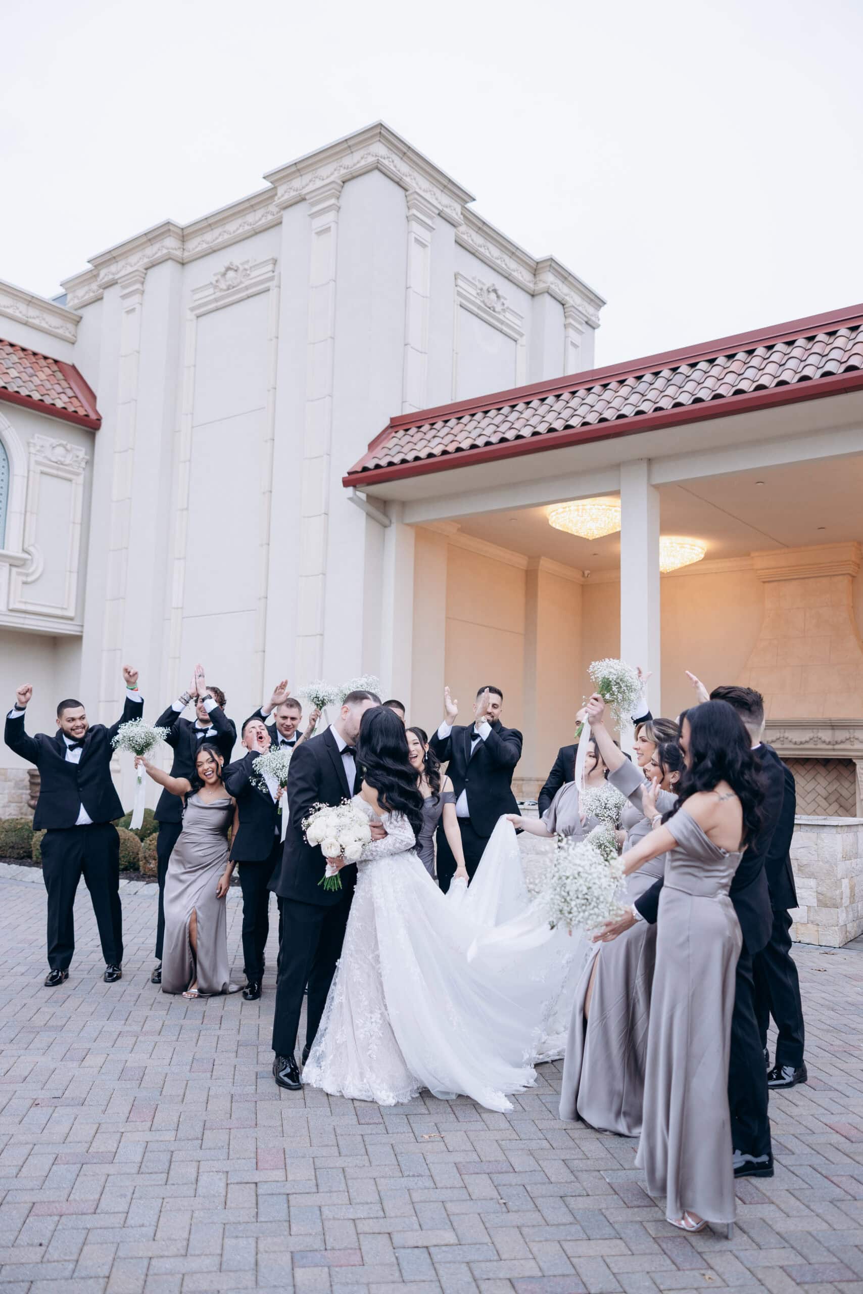 Bride and groom kissing outdoors with wedding party cheering and holding bouquets, in front of a sophisticated building with tiled roof and ornate columns.