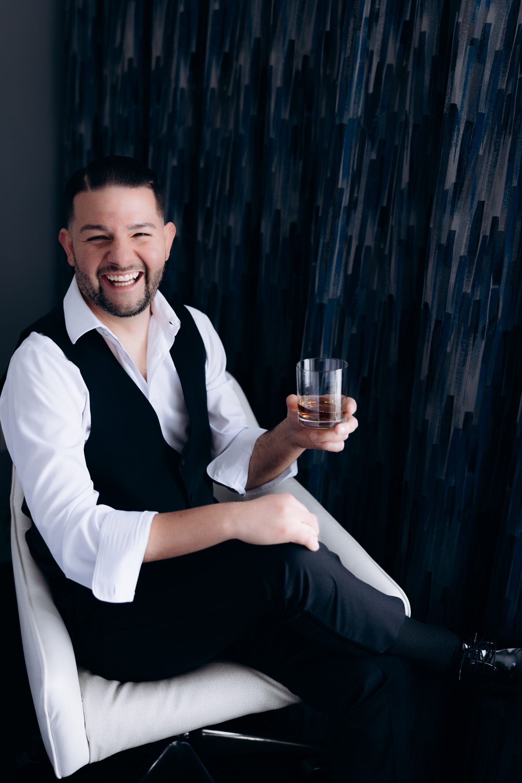 Man wearing white shirt and black vest seated on a chair holding a glass of whiskey, smiling broadly against a dark abstract patterned wall.