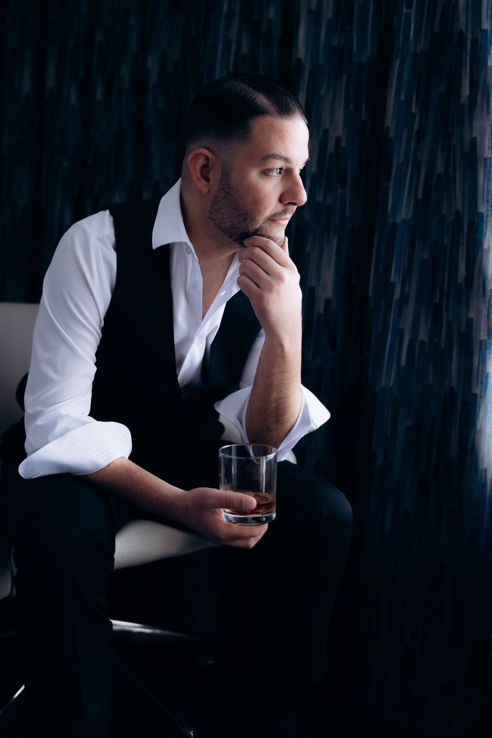 Man in white dress shirt and black vest seated, holding a glass of whiskey, looking contemplatively to the side against a dark textured background.