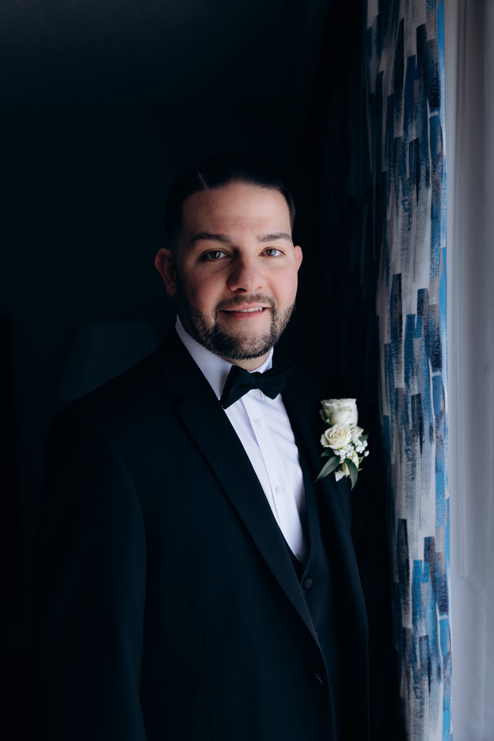 Portrait of a groom in black tuxedo and bow tie standing near a window with patterned blue and white curtains, softly lit against a dark background.