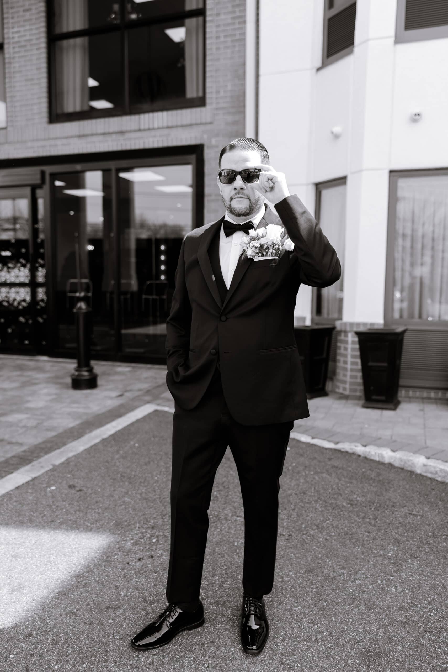 Groom in black tuxedo with bow tie and white boutonniere adjusting sunglasses outside modern building entrance at The Grand.