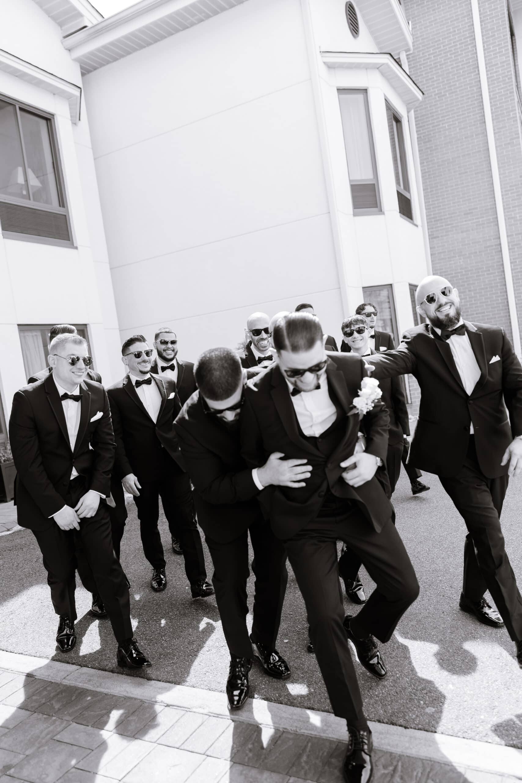Group of groomsmen in black tuxedos and sunglasses sharing a joyful moment outdoors near white and brick buildings at The Grand.