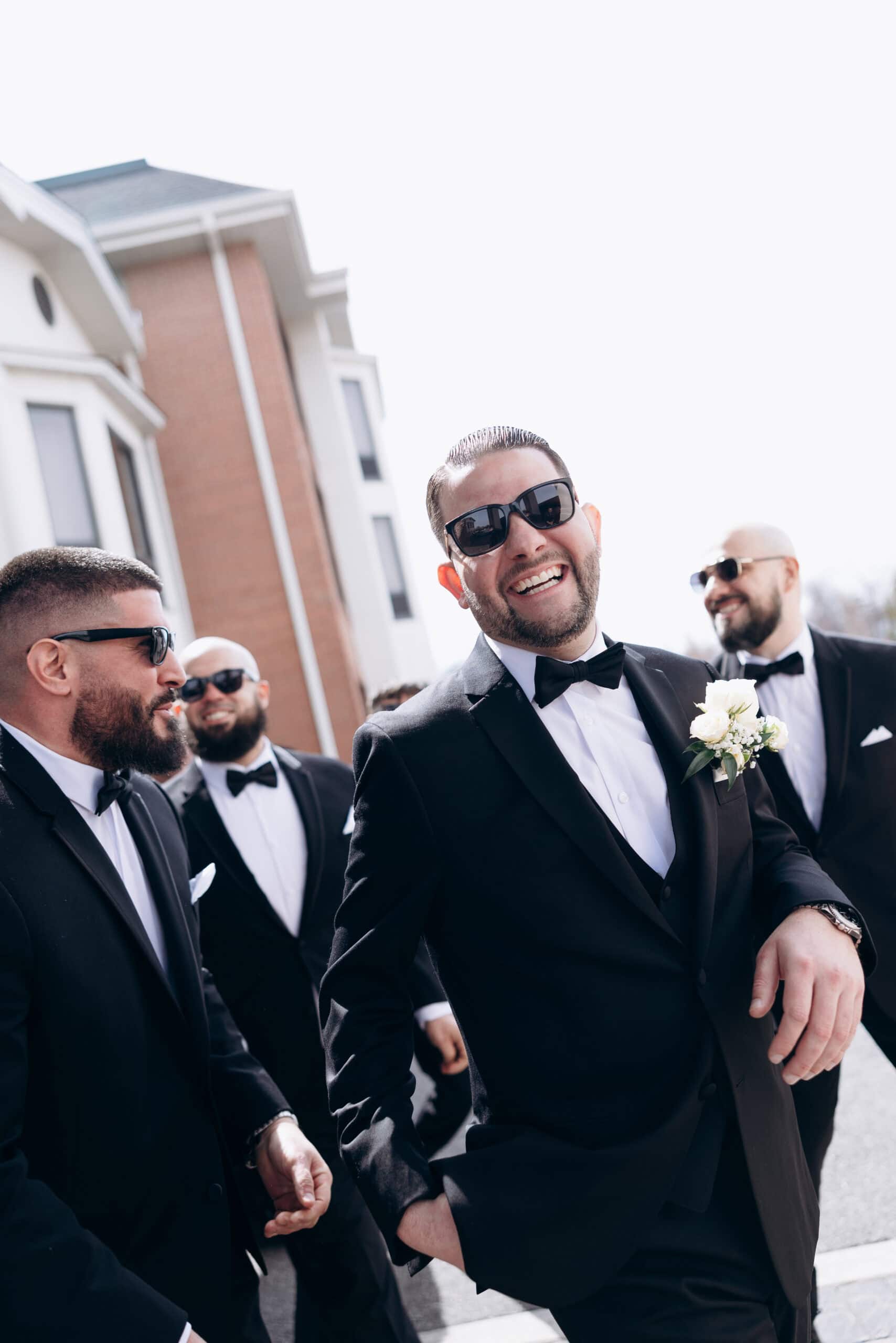 Groom and groomsmen in black tuxedos and sunglasses laughing outside near a brick and white building, capturing a joyful wedding moment.