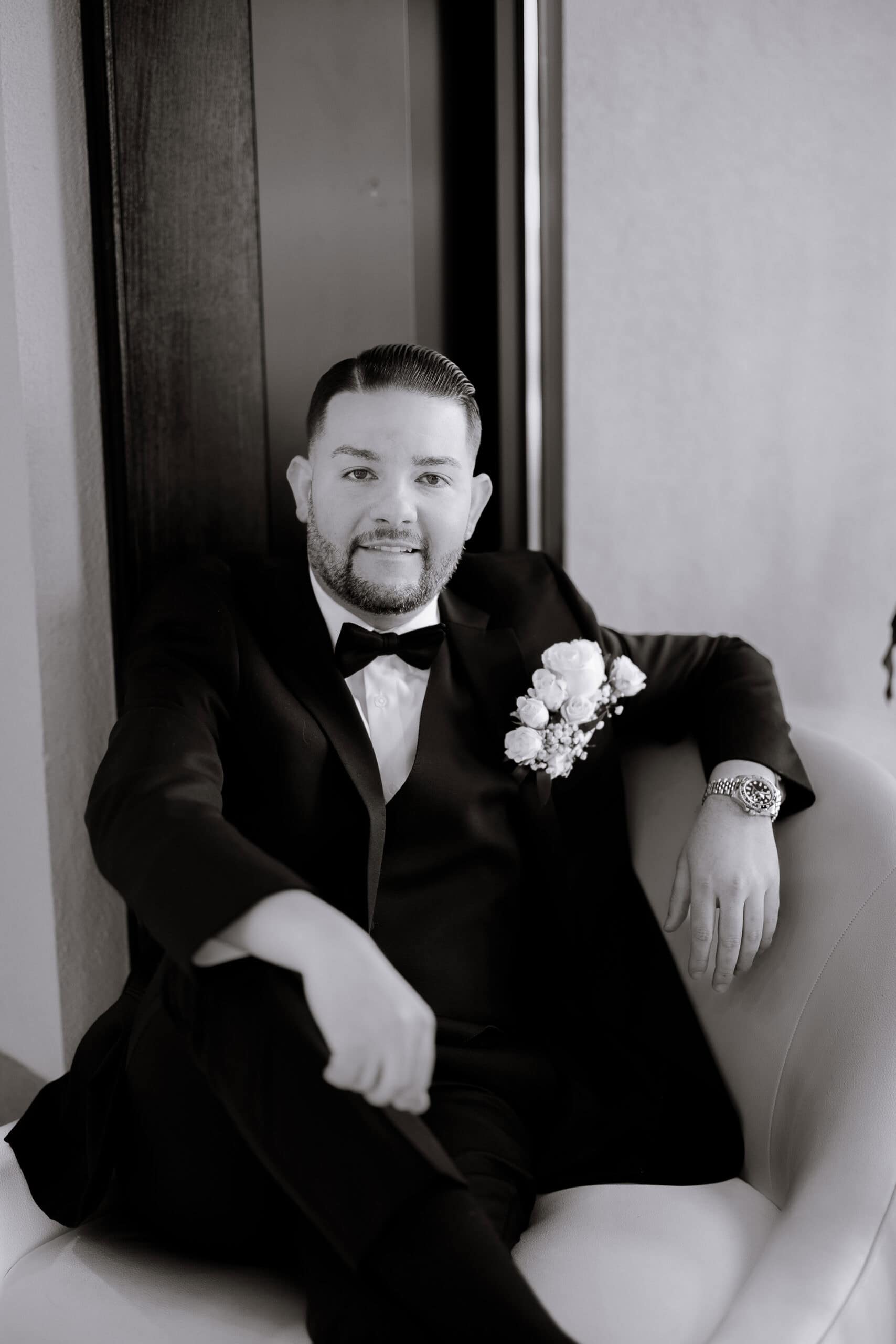 Black and white portrait of a groom seated in a modern chair wearing a black tuxedo and bow tie, with a white rose boutonniere and a luxury watch.