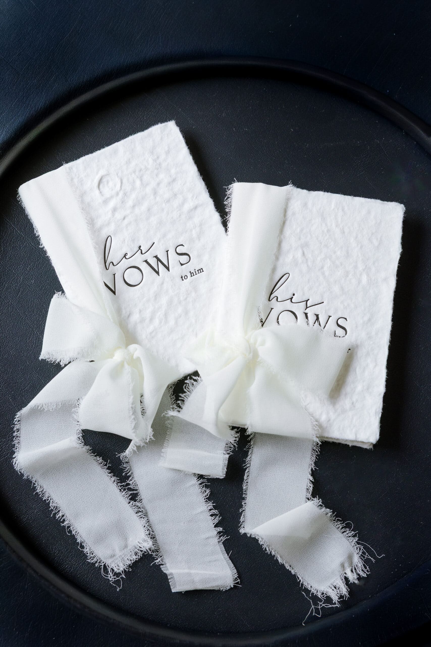 Close-up of two white textured vow books labeled 'her vows to him' and 'his vows to her' tied with soft white silk ribbons on a black circular tray.
