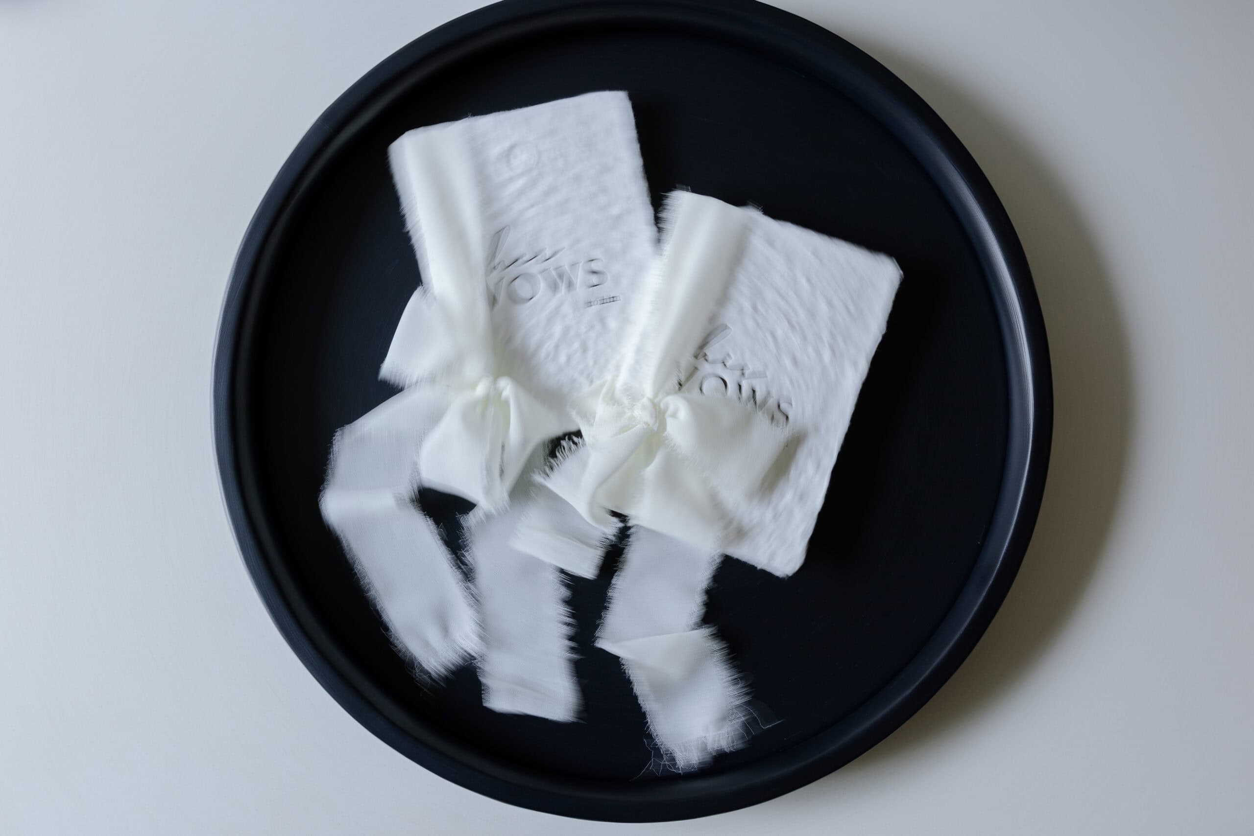 Two white textured vow books with frayed edges and ribbon ties placed on a round black tray on a white surface.