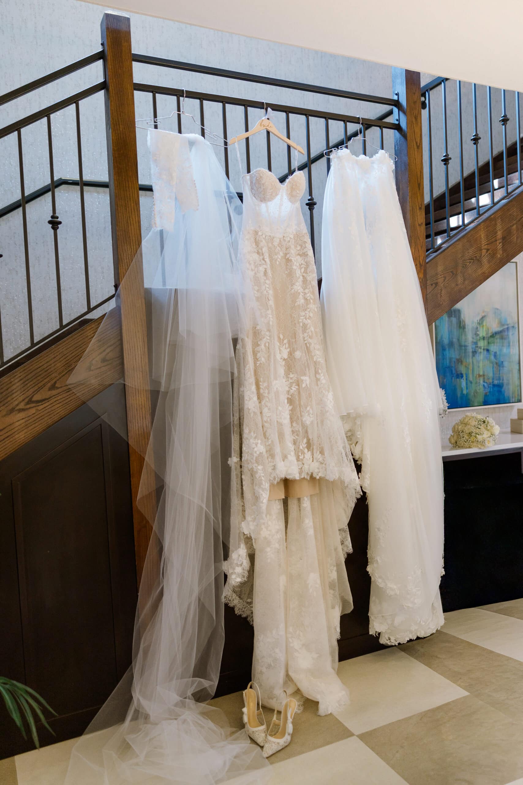 A lace wedding dress and long veil hanging on a wooden staircase railing with a pair of embroidered bridal heels on a checkered floor beneath.