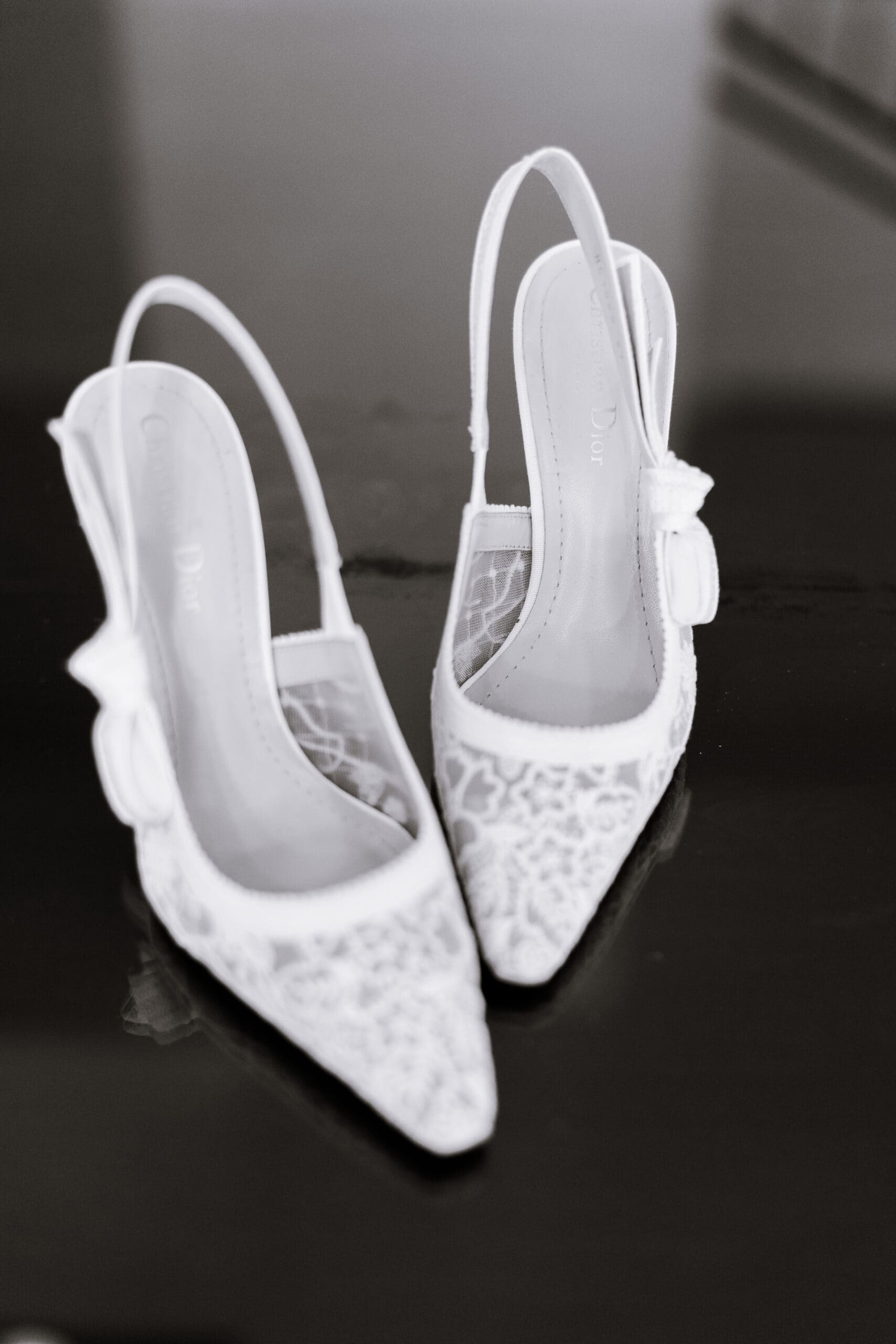Close-up of white Dior lace slingback bridal heels with bows on a reflective dark surface.