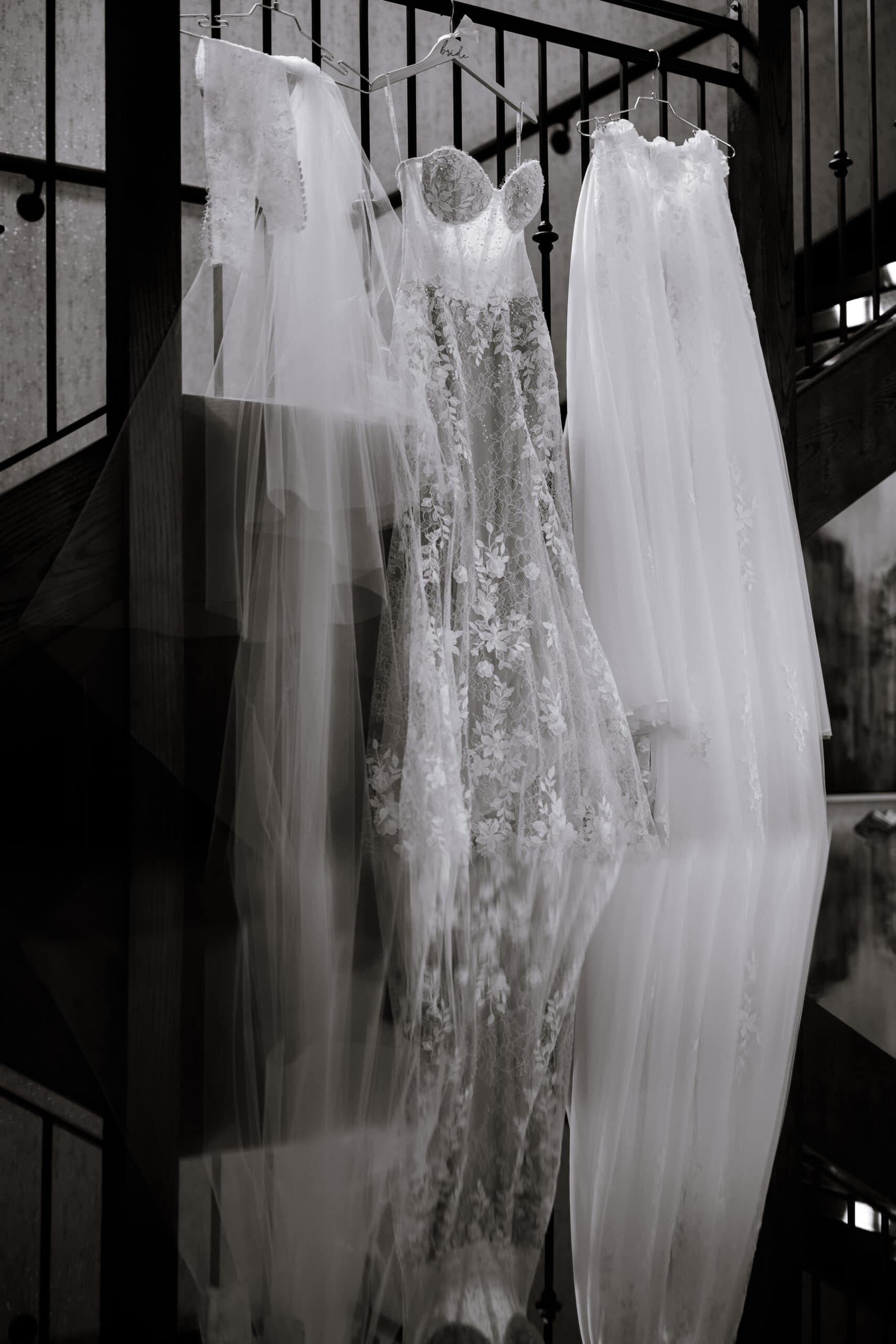 Close-up of a lace wedding dress and veil hanging on a decorative hanger labeled 'bride' against a dark metal railing, showcasing intricate floral embroidery and sheer fabric.
