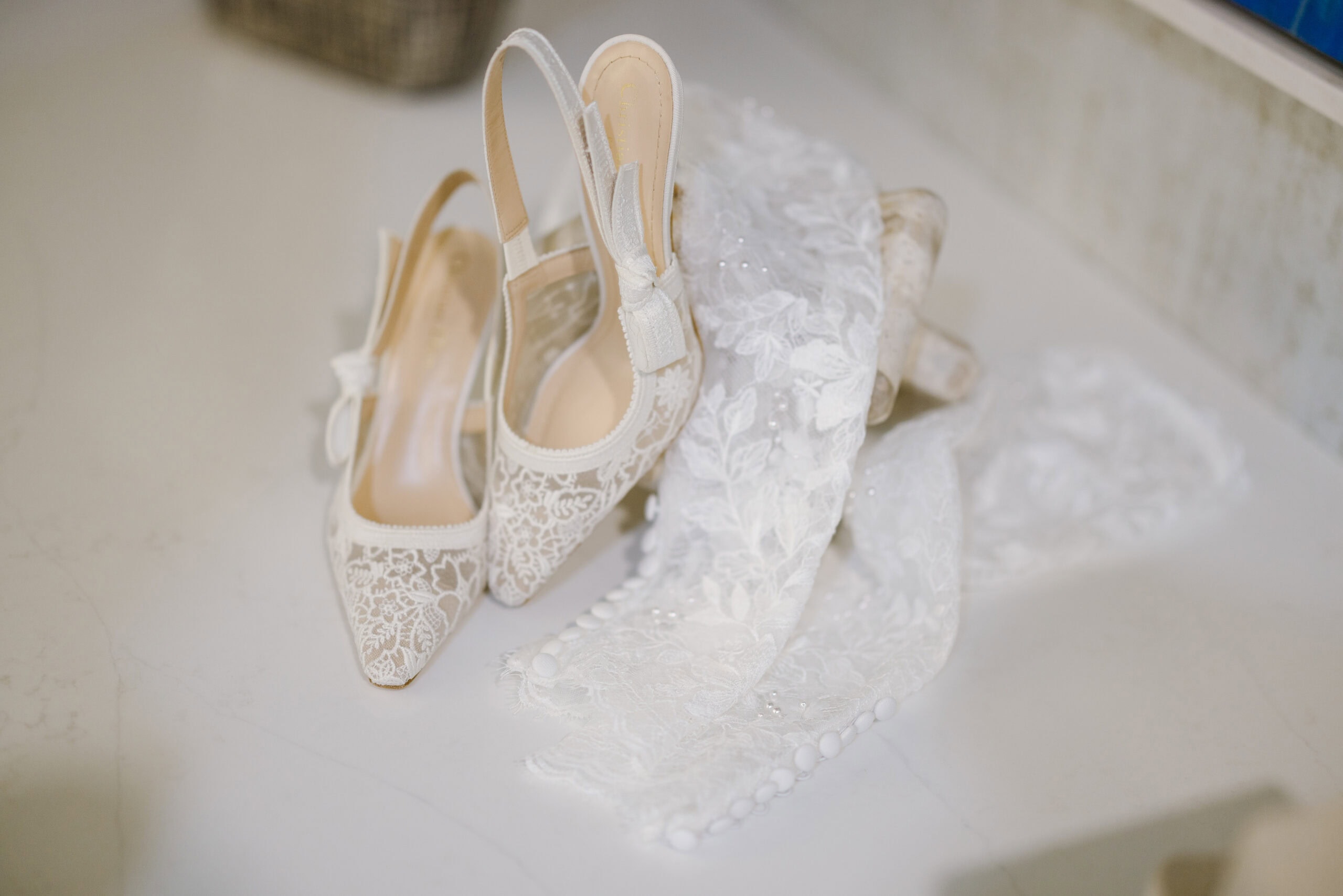 Close-up of white lace bridal slingback shoes next to a lace veil with buttons and bead details on a marble surface.
