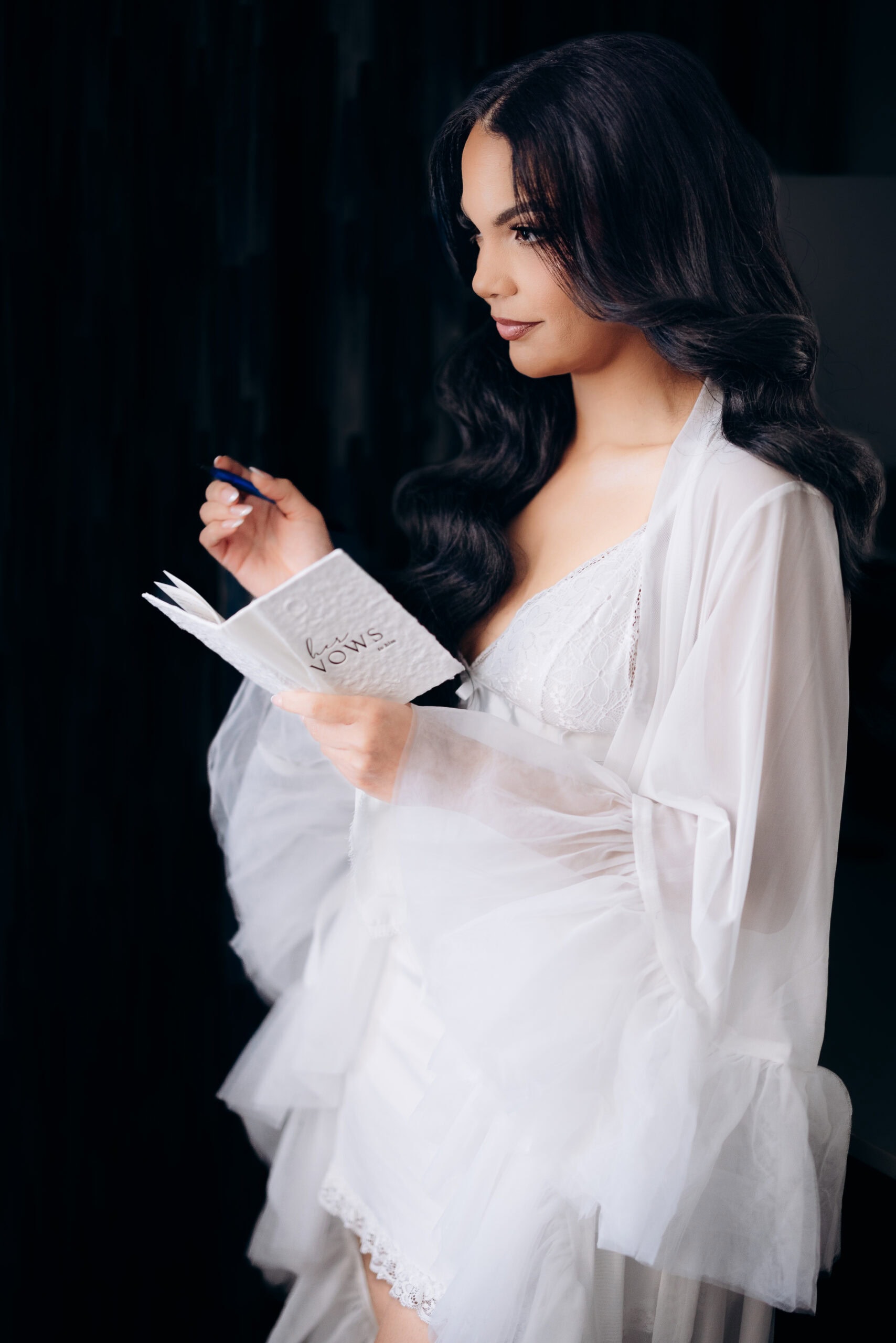 Side profile of a bride with long wavy hair wearing a sheer white robe over lace lingerie, holding a pen and embossed leather vow booklet against a dark background.