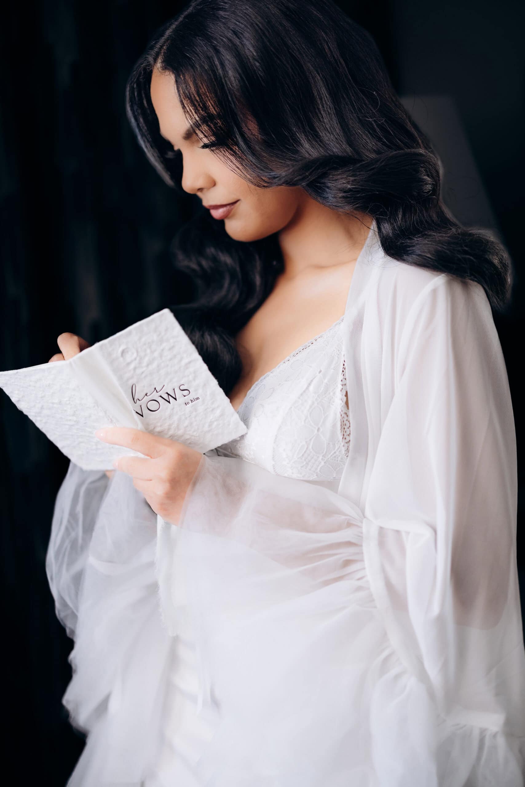 Close-up of a bride with dark wavy hair wearing a white lace dress and sheer robe, reading a textured vow booklet titled 'her vows to him' against a dark background.
