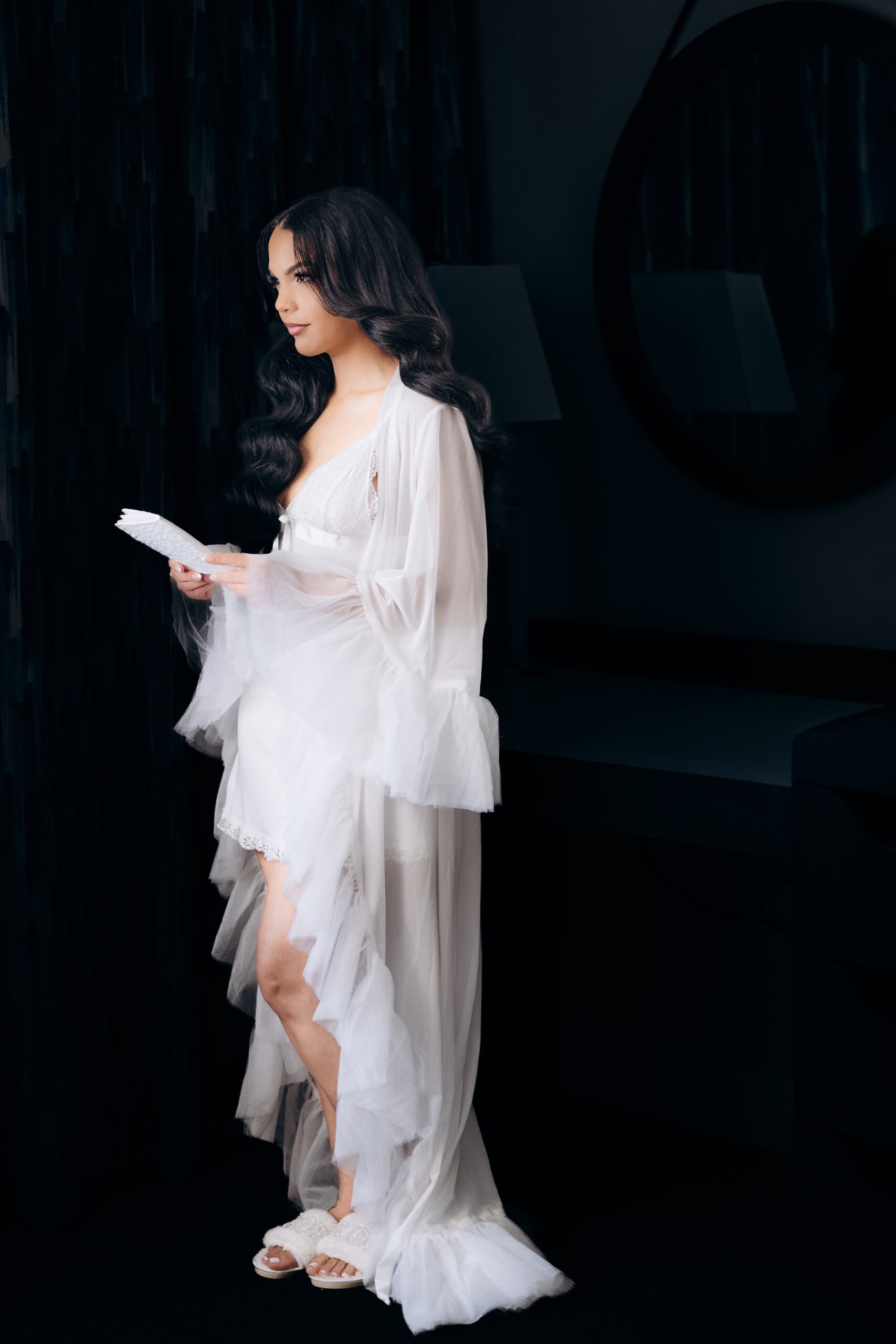 Bride standing in a sheer white robe and lace lingerie, holding a folded letter, illuminated softly against a dark room with a round mirror and lamp in the background.
