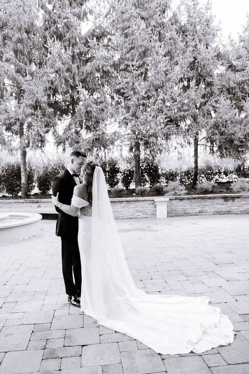 Bride and groom in an intimate embrace on a stone-paved courtyard, surrounded by tall evergreen trees, bride’s long veil trailing on the ground.
