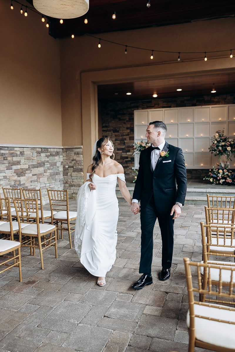 Bride in off-shoulder white gown and groom in black tuxedo holding hands walking through aisle with gold chairs and stone wall at The Stone House, Warren NJ.