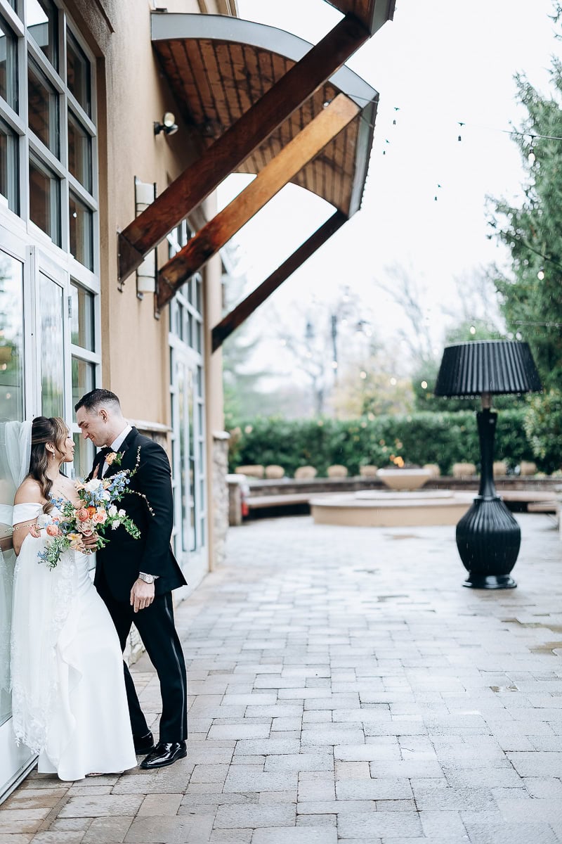 Bride and groom leaning close together beside glass doors under wooden awnings outside The Stone House, Warren, NJ, with soft natural light and greenery in background.