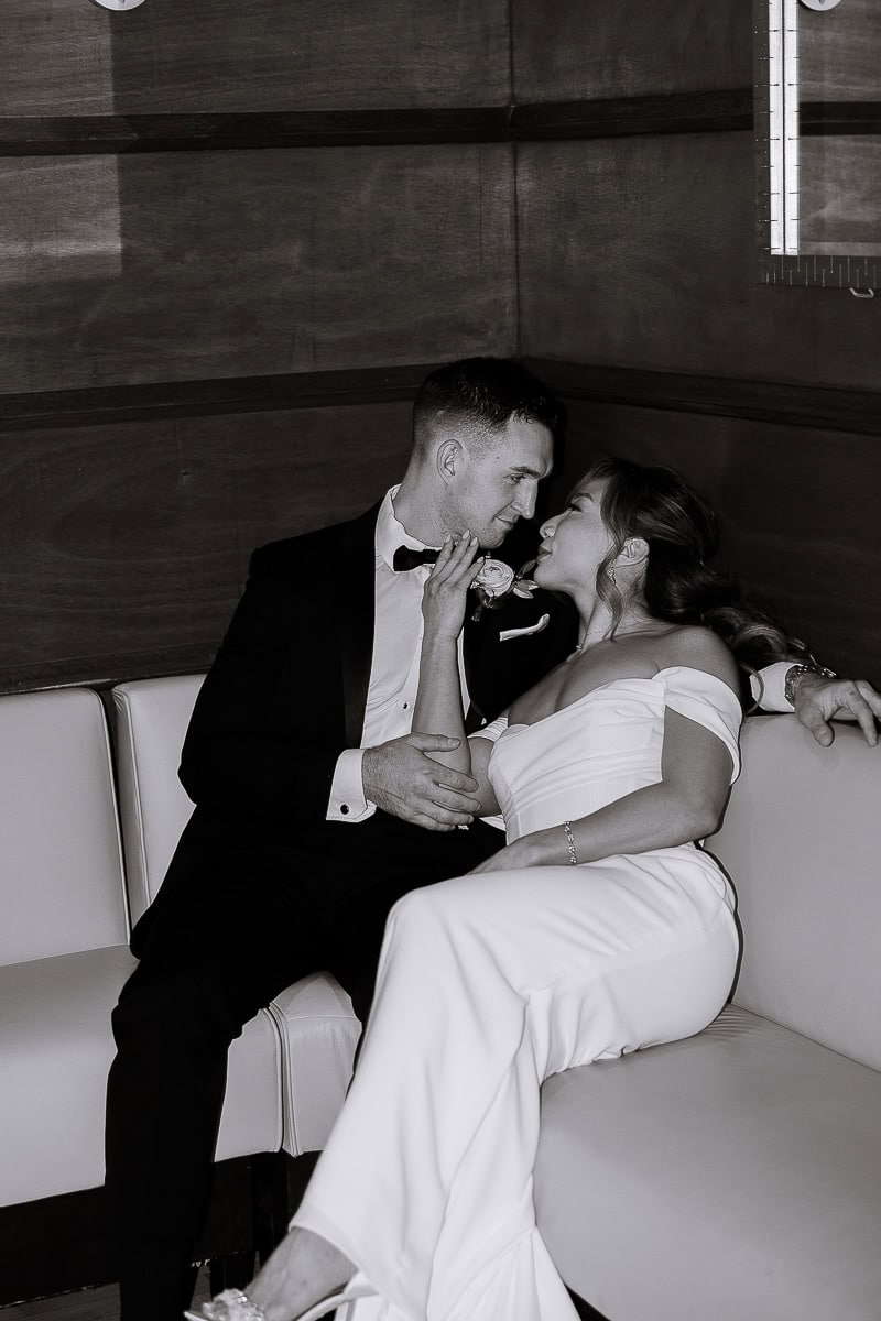 Bride in off-shoulder white gown and groom in black tuxedo seated closely on leather bench, sharing an intimate gaze indoors at The Stone House, Warren, NJ.