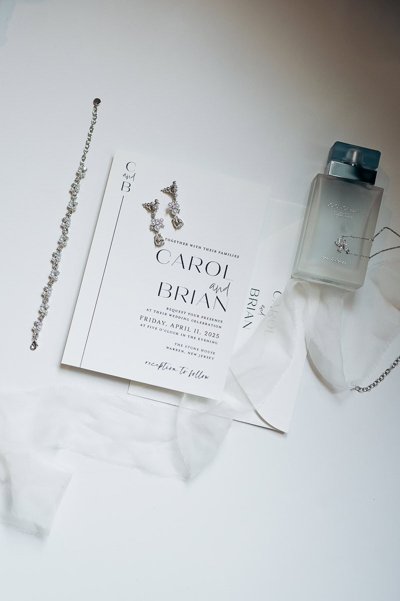 Flat lay of a minimalist wedding invitation for Carol and Brian at The Stone House, Warren, NJ, surrounded by crystal earrings, bracelet, sheer fabric, and a frosted glass perfume bottle.