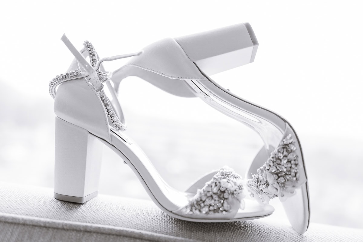 Pair of white satin bridal heels with crystal embellishments and ankle straps, resting on a textured surface with soft background light.