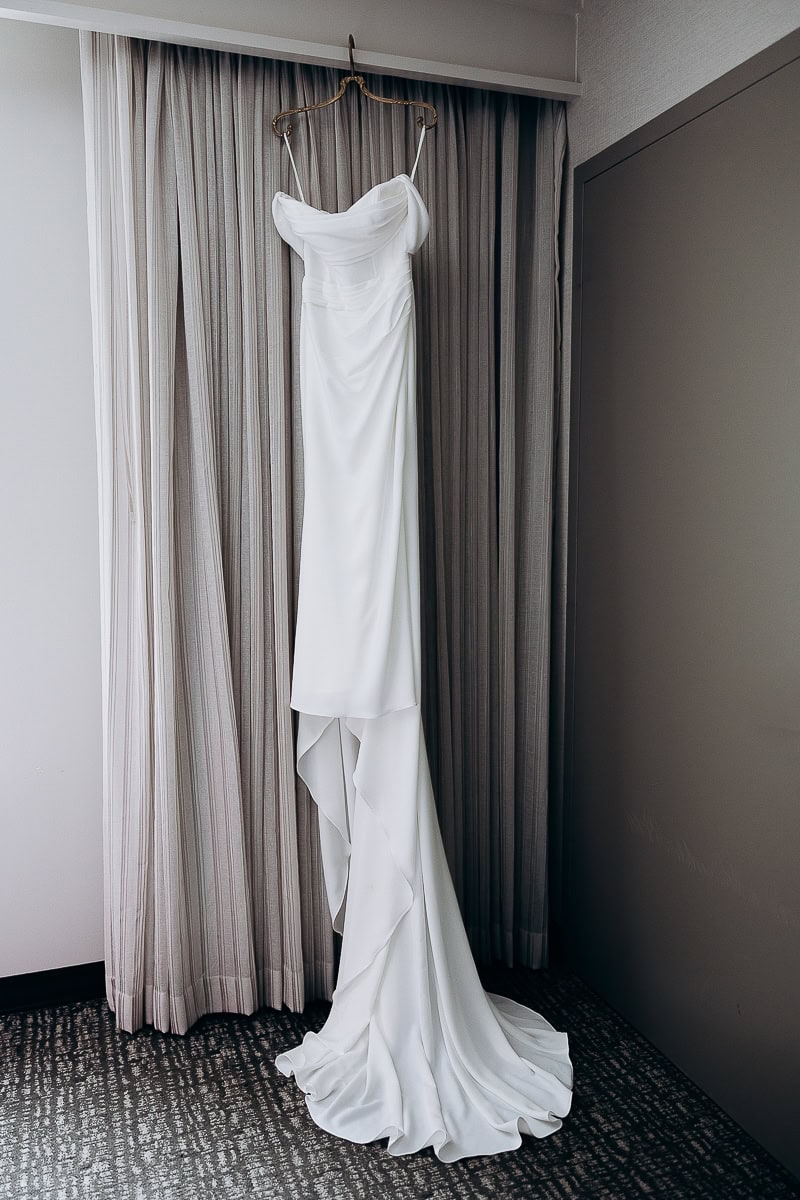 White minimalist wedding dress with draped bodice and flowing train hanging on hanger in front of neutral curtains at The Stone House, Warren, NJ.