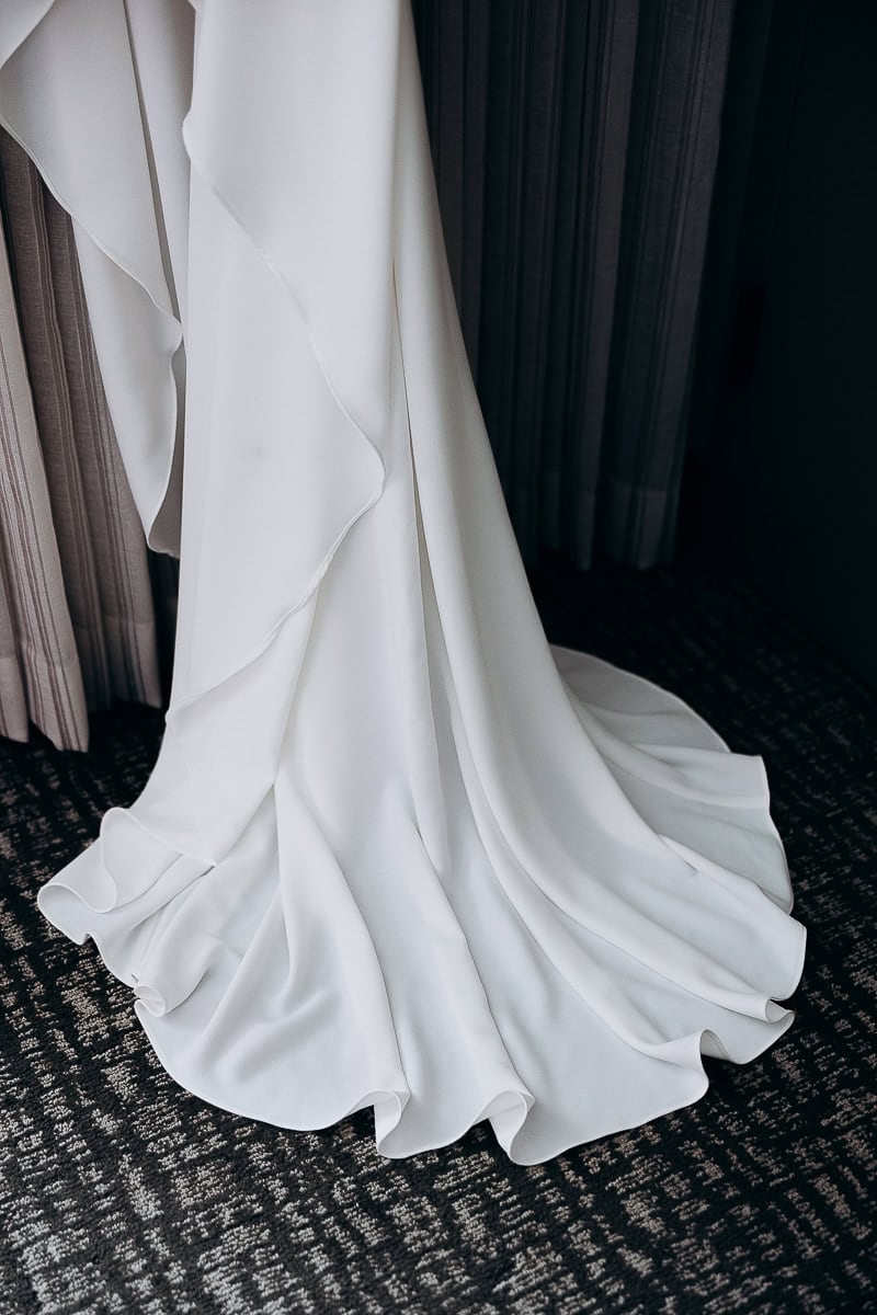 Close-up of the flowing, layered hem of a minimalist white wedding gown on a dark patterned carpet near curtains.