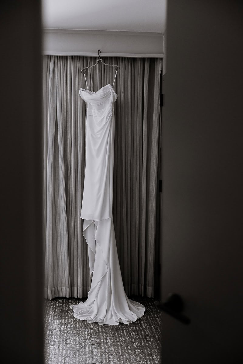 White wedding gown hanging on a hanger in front of pleated curtains, viewed through a partially open door at The Stone House, Warren, NJ.