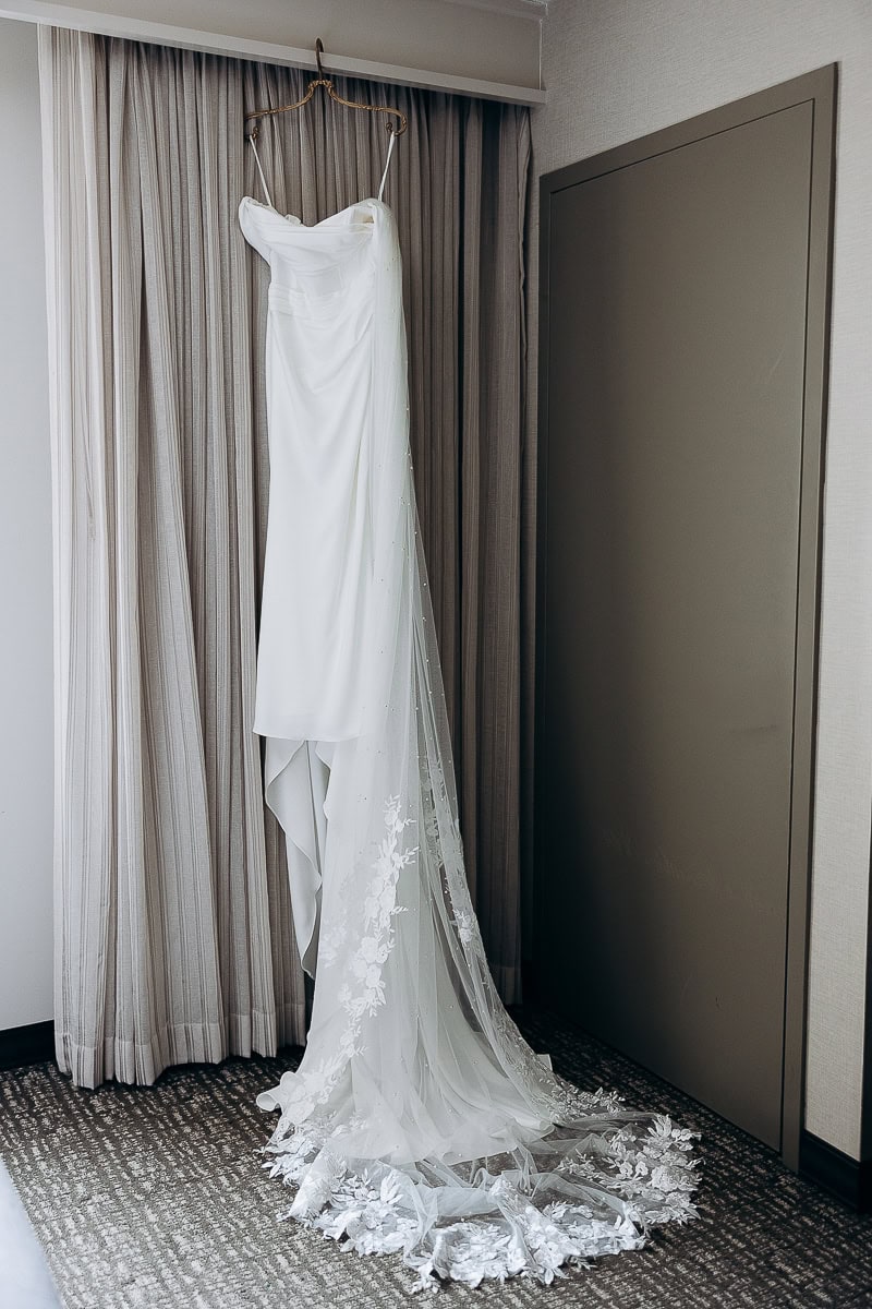 White satin wedding dress with a long floral lace veil hanging on a decorative hanger in a softly lit room with neutral curtains and carpet.