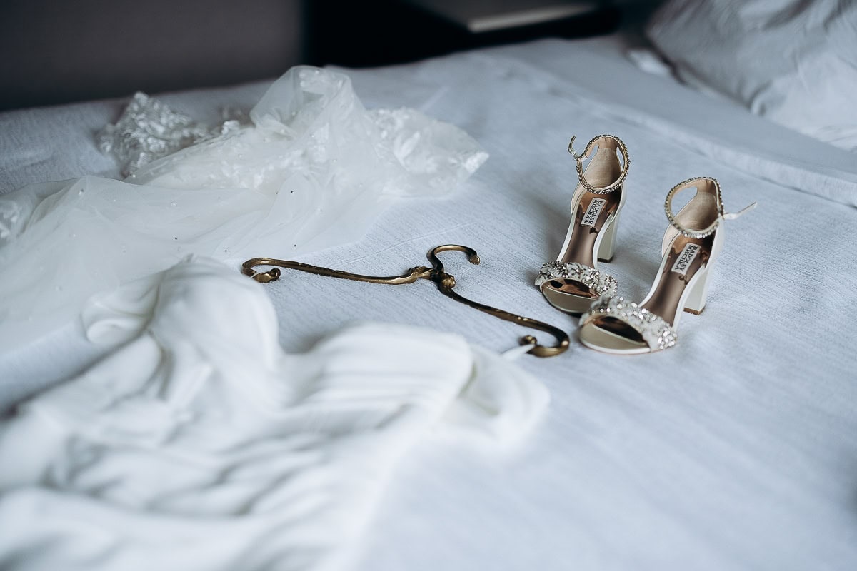 Embellished bridal heels and lace wedding gown laid out on a white bedspread with a decorative hanger at The Stone House, Warren, NJ.
