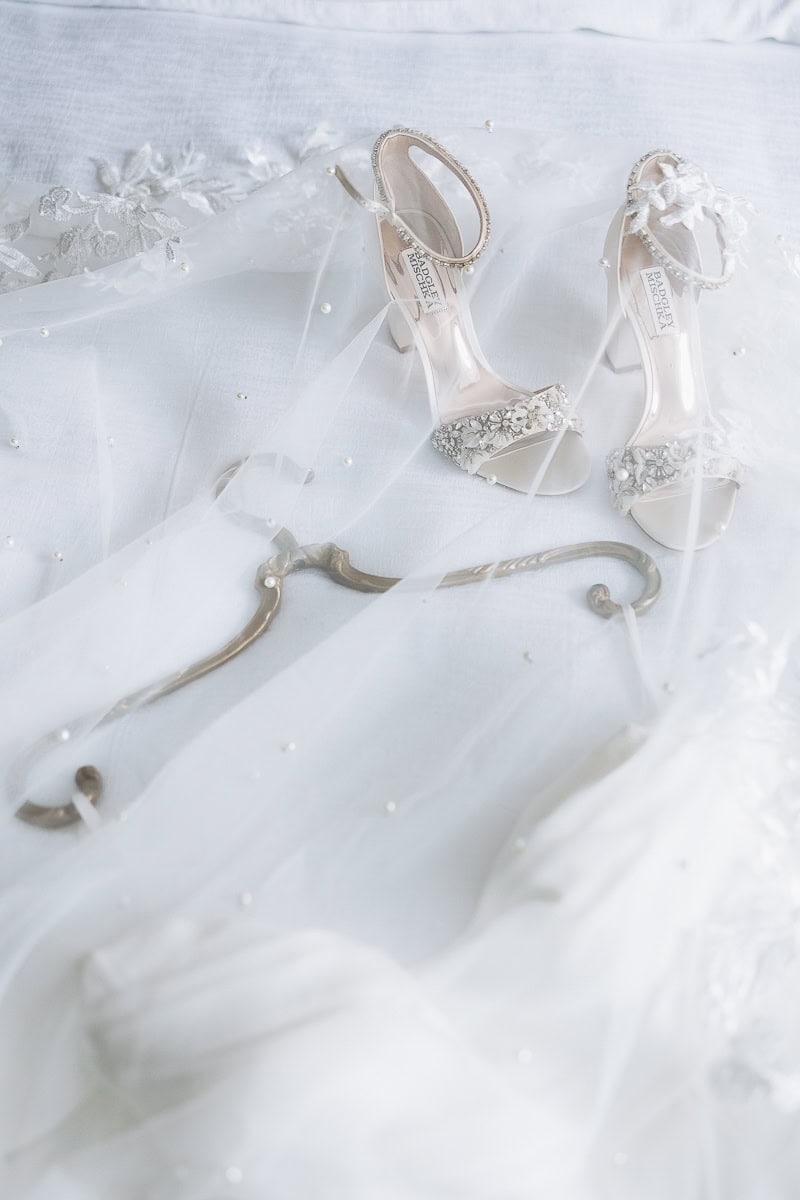 Pair of embellished Badgley Mischka bridal heels on a white bed draped with a pearl-adorned veil and lace details.