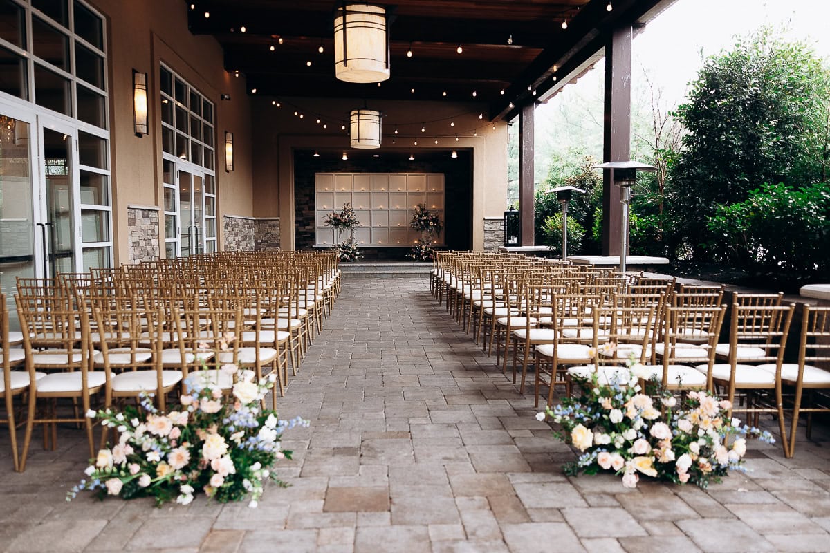 Covered outdoor wedding ceremony setup with rows of gold chiavari chairs, floral arrangements at aisle entrance, and warm lantern lighting at The Stone House, Warren NJ.