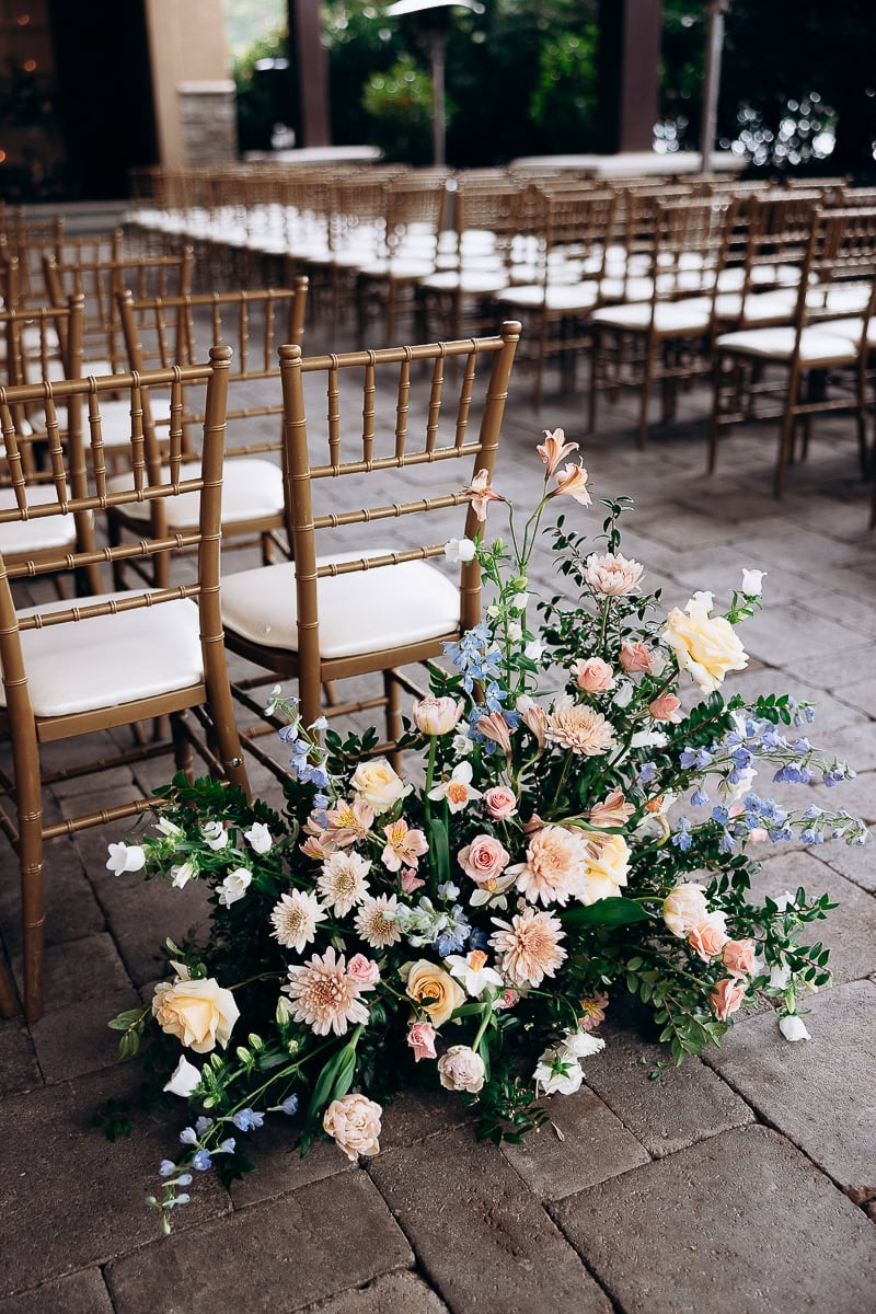Pastel floral arrangement with roses, dahlias, and delphiniums beside gold chiavari chairs on stone floor at wedding venue.