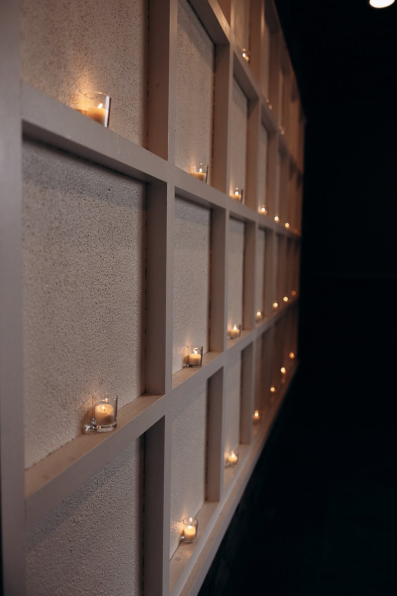Close-up of votive candles in glass holders placed in a grid-patterned wall niche, softly glowing in dim light at The Stone House, Warren, NJ.